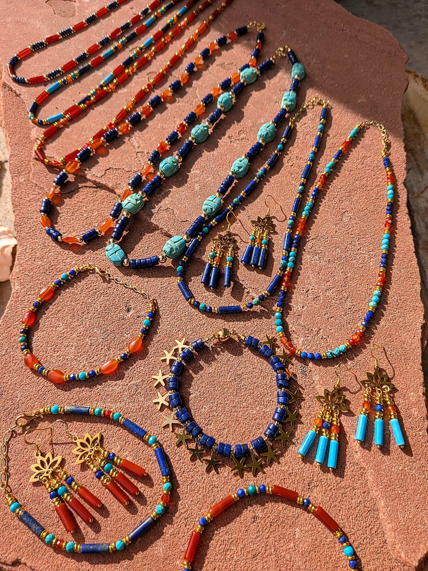 A collection of handmade beaded jewelry, including necklaces, bracelets, and earrings, displayed on a textured terracotta surface. The jewelry features vibrant blue, orange, and turquoise beads with gold-toned metal charms and tassels.