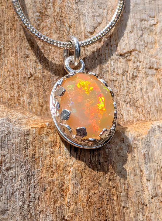 A silver chain necklace with an oval pendant featuring a warm orange and yellow opal-like gemstone in a textured silver bezel, displayed on a rustic wood surface.