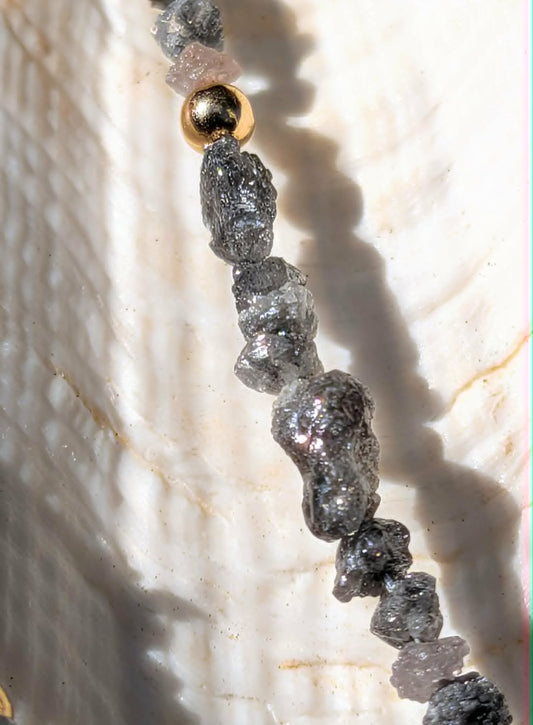 A close-up of a beaded strand featuring rough, dark gray to black rough uncut diamond nugget beads and a single metallic gold bead, resting on a textured white clam shell.