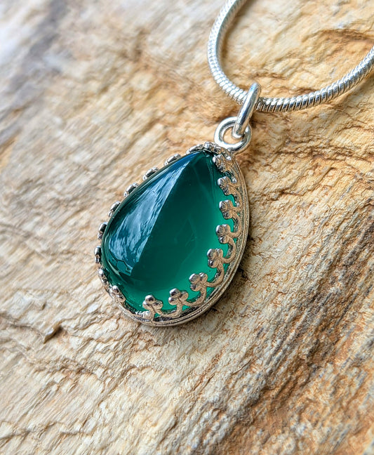 A teardrop-shaped green gemstone pendant with a silver filigree border and a silver snake chain, displayed on a textured wood background.