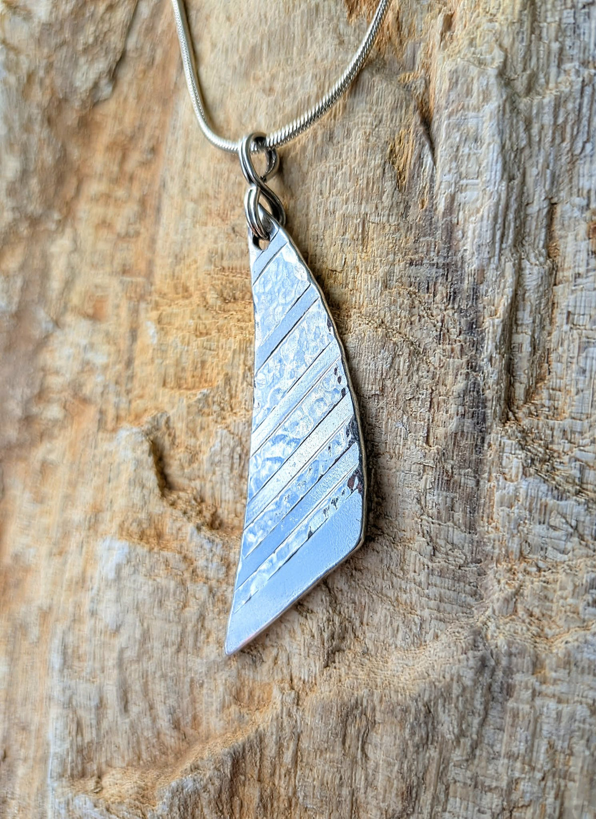 A silver sail-shaped pendant necklace with diagonal textured stripes, hanging from a silver snake chain against a rustic, textured wood background.