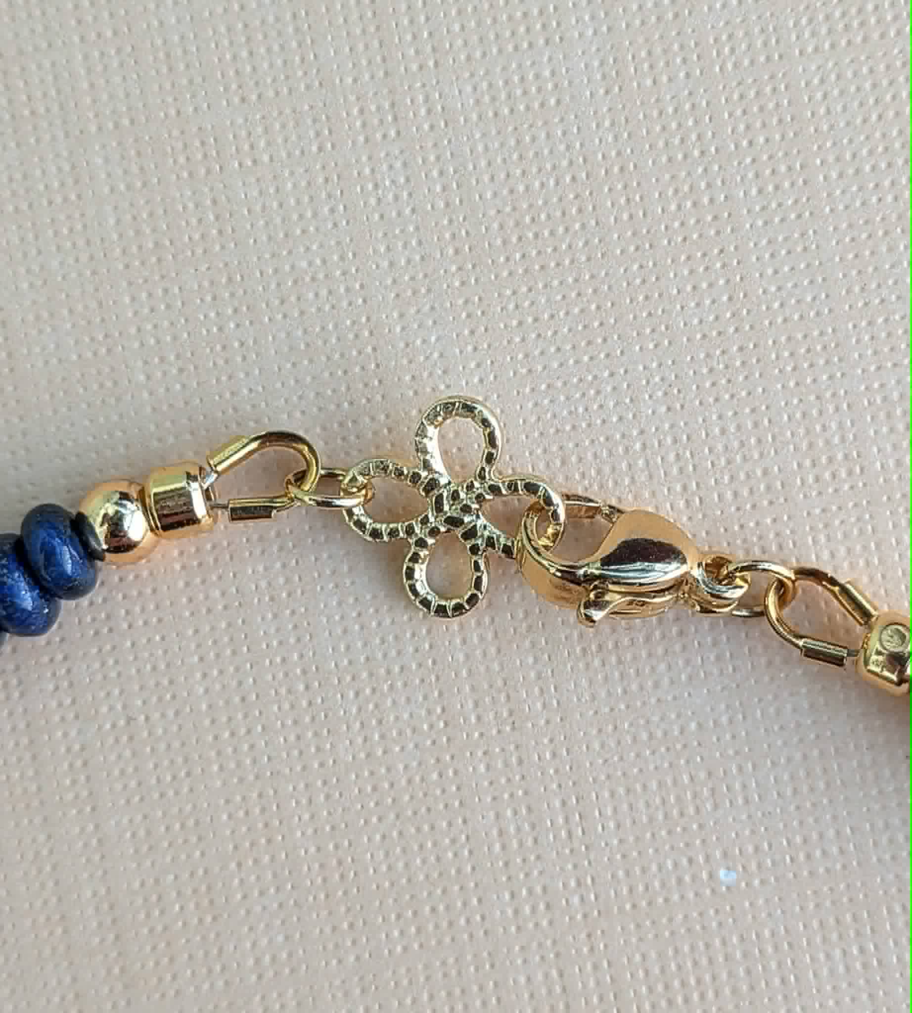 Close-up of a gold decorative clover-shaped charm, lobster clasp, and blue rondelle beads on a textured beige background.