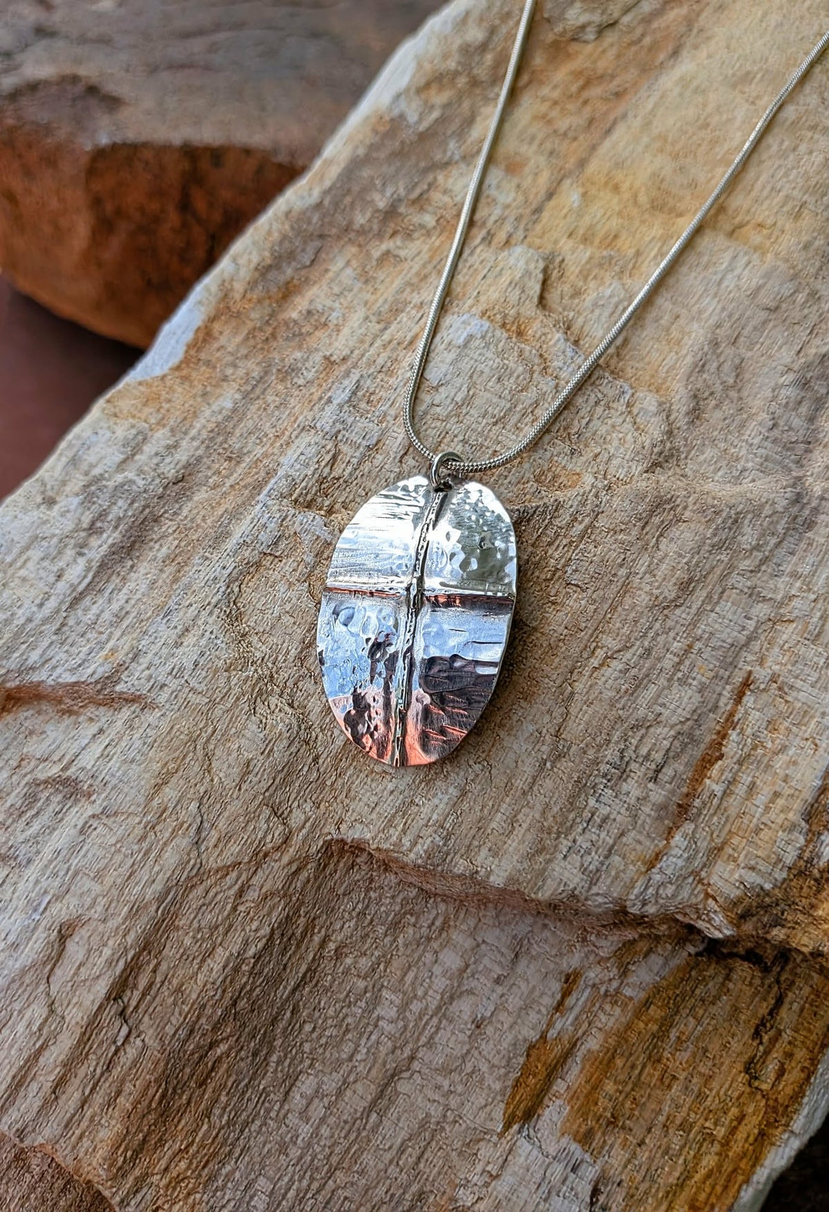 A silver shield-shaped pendant necklace with a textured finish, resting on a rustic piece of wood.