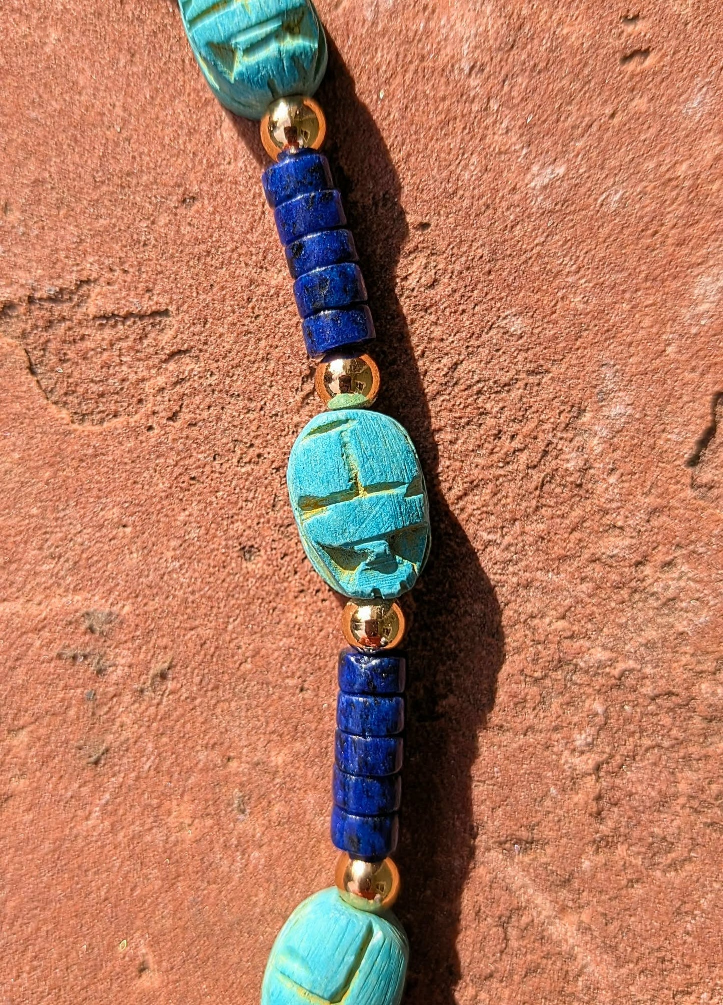 A close-up of a beaded necklace featuring turquoise scarab beetle charms, deep blue cylindrical beads, and small gold spacer beads, laid diagonally on a textured terracotta surface.