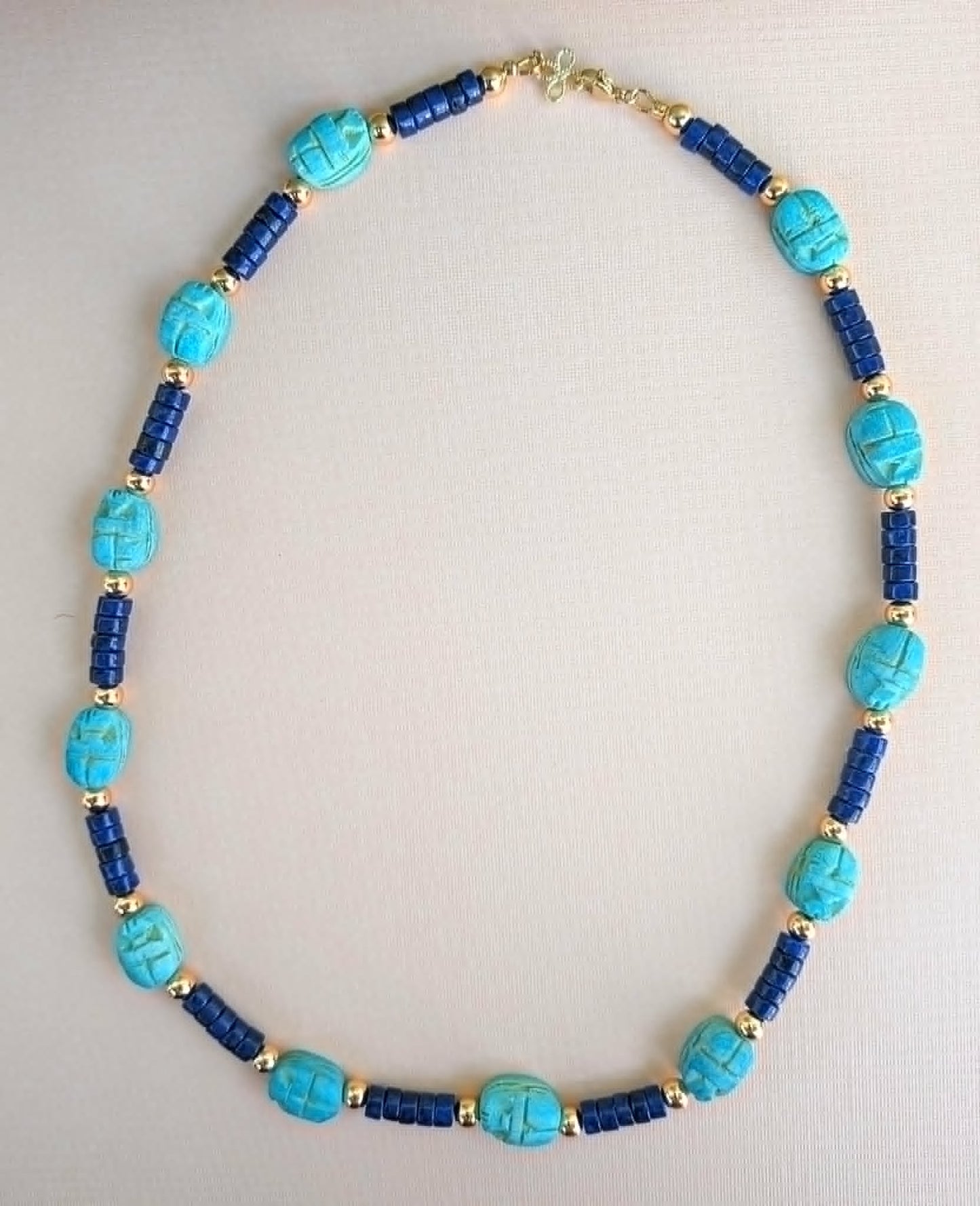A beaded necklace featuring turquoise and dark blue beads separated by small gold spacer beads, laid out on a light beige background.