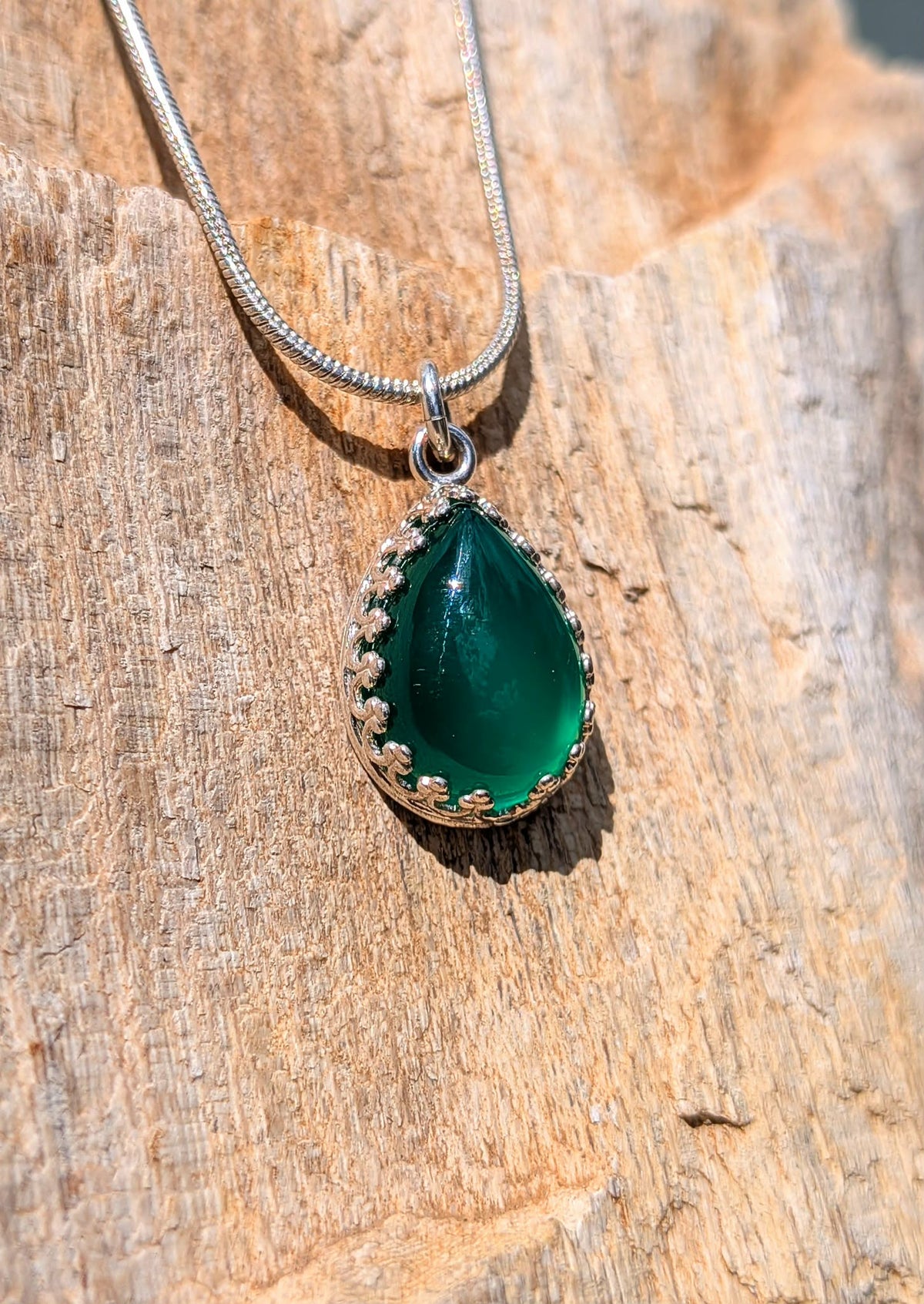 A silver teardrop pendant necklace with a green stone and small gold floral accents, resting on a textured wooden surface.