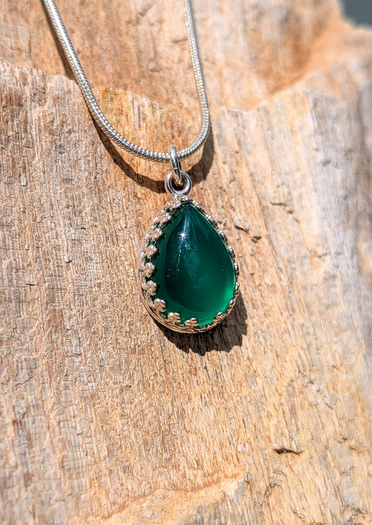 A silver teardrop pendant necklace with a green stone and small gold floral accents, resting on a textured wooden surface.
