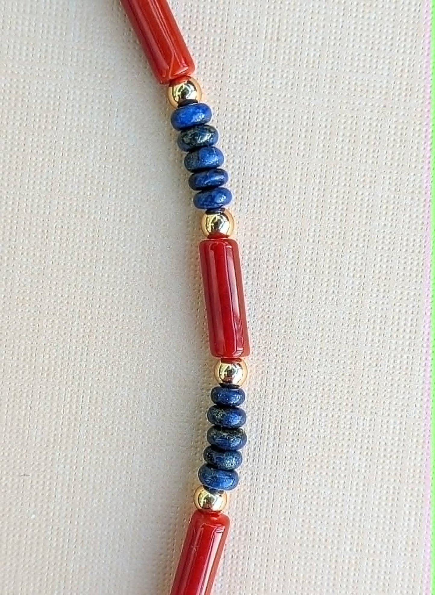 A close-up of a beaded necklace featuring polished red cylindrical beads, dark blue round beads, and small metallic gold spacer beads, laid flat on a light beige textured fabric background.