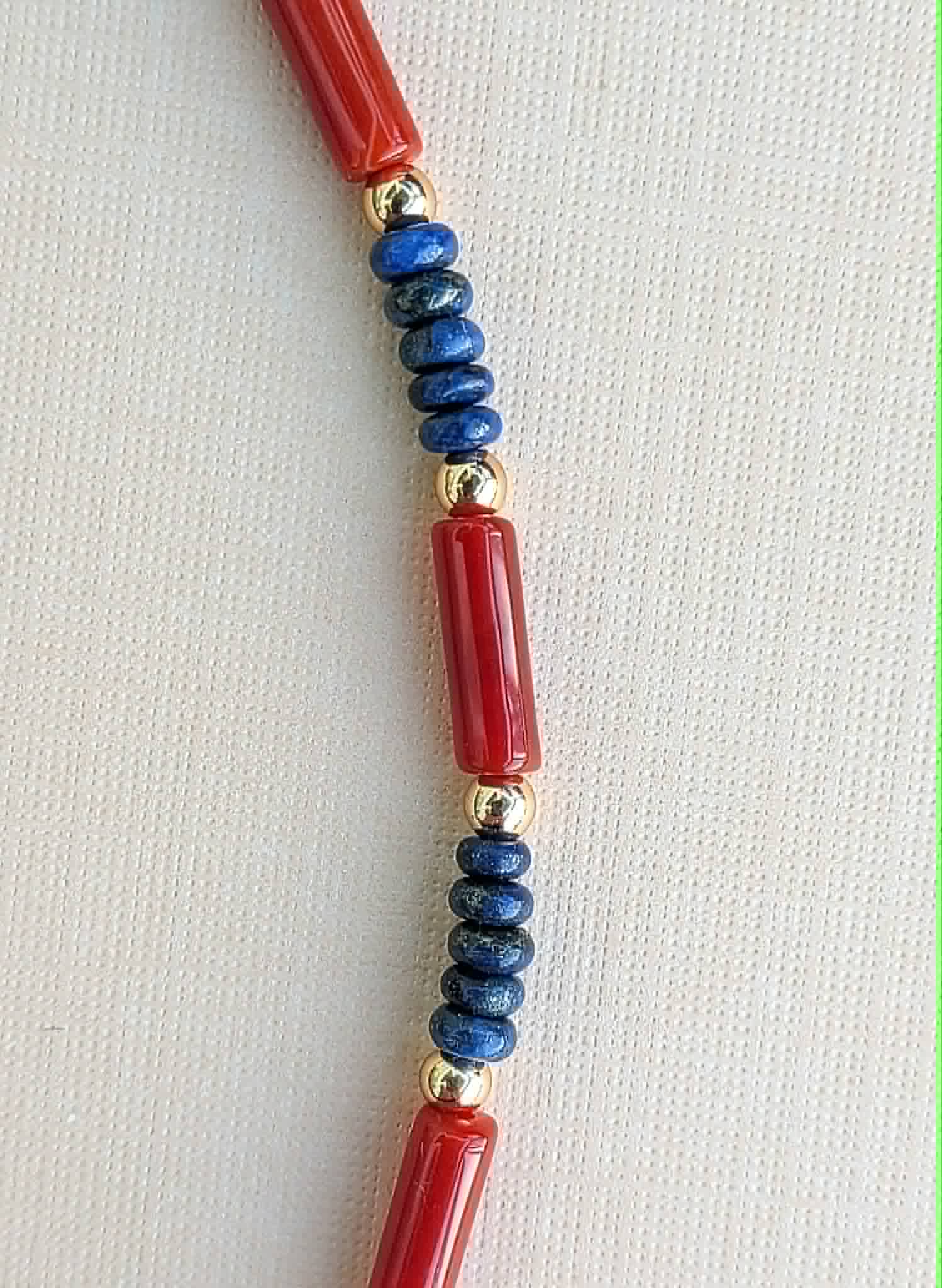 A close-up of a beaded necklace featuring polished red cylindrical beads, dark blue round beads, and small metallic gold spacer beads, laid flat on a light beige textured fabric background.