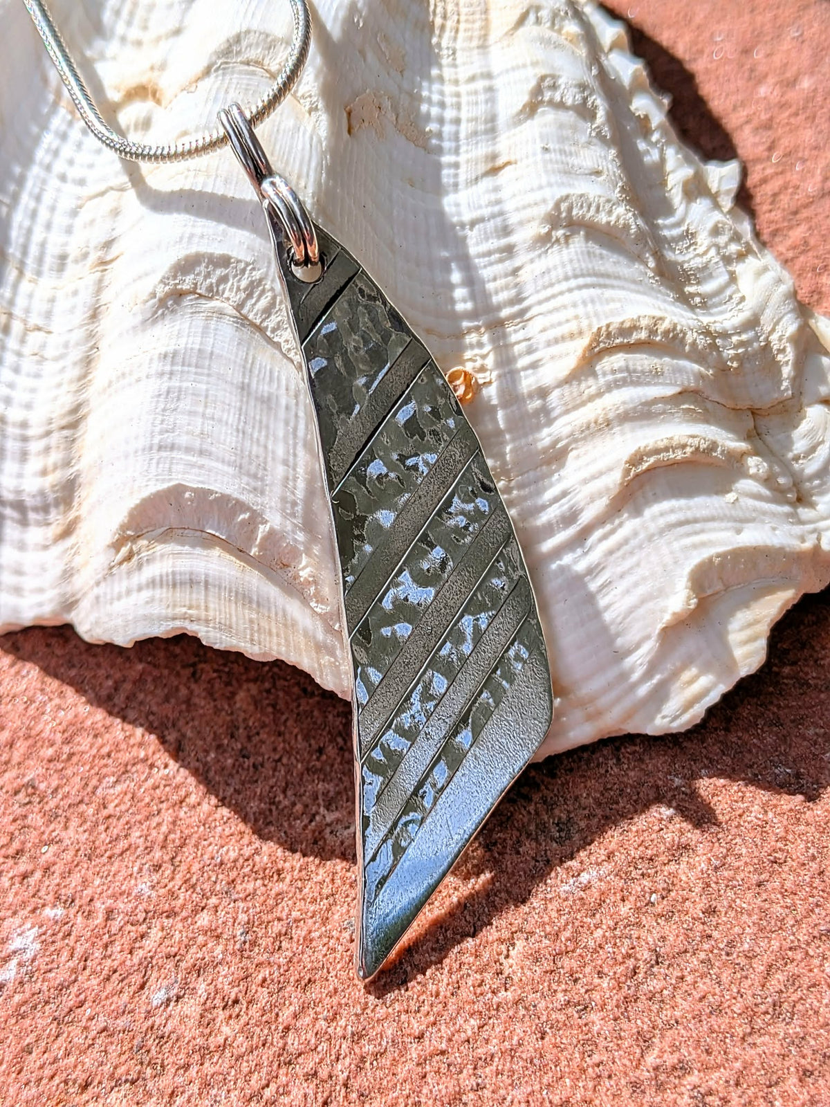 A silver metal pendant necklace with a textured, hammered finish and diagonal ridges, resting on a large white seashell over a reddish-brown sandy surface.