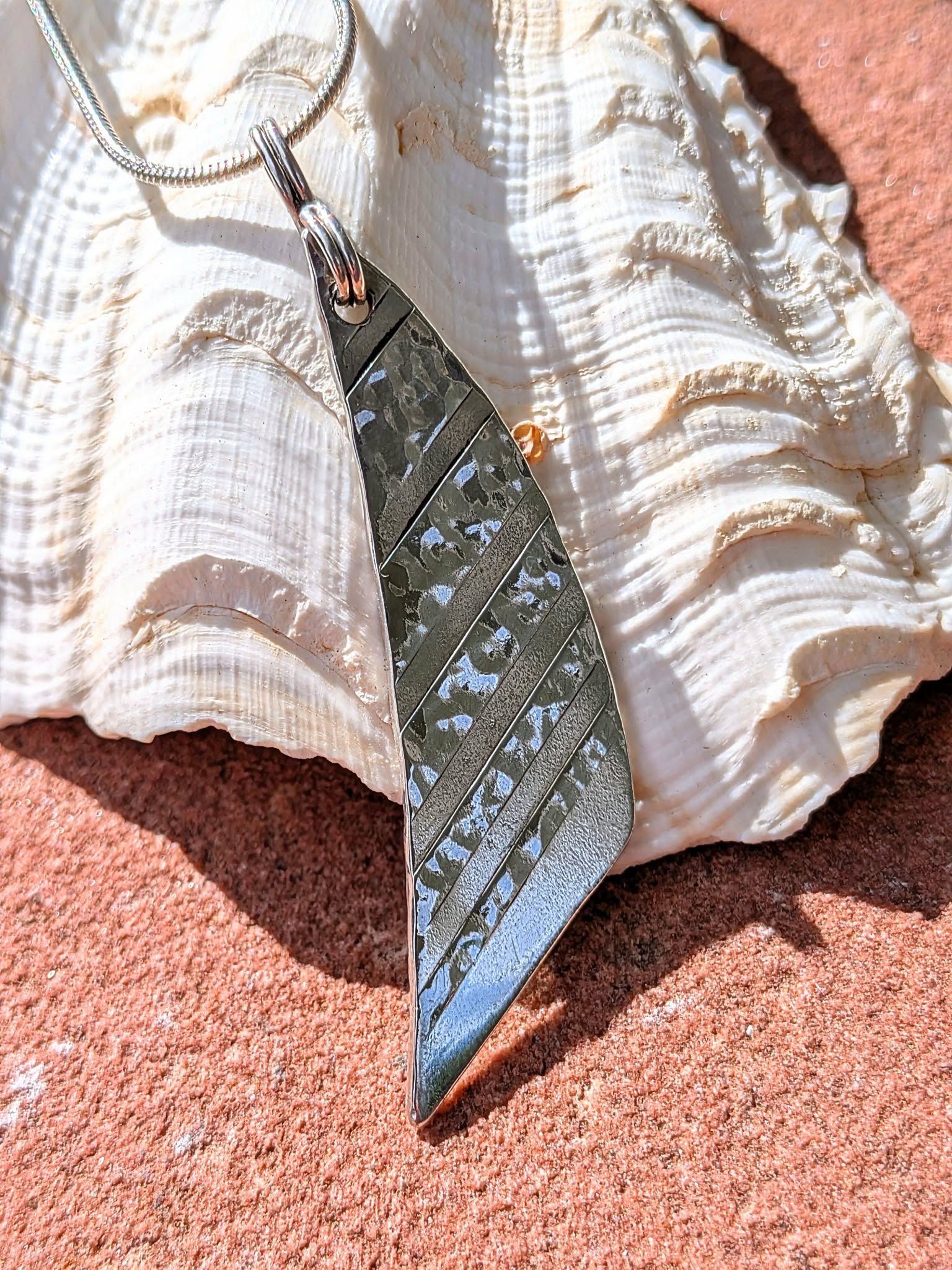 A silver metal pendant necklace with a textured, hammered finish and diagonal ridges, resting on a large white seashell over a reddish-brown sandy surface.