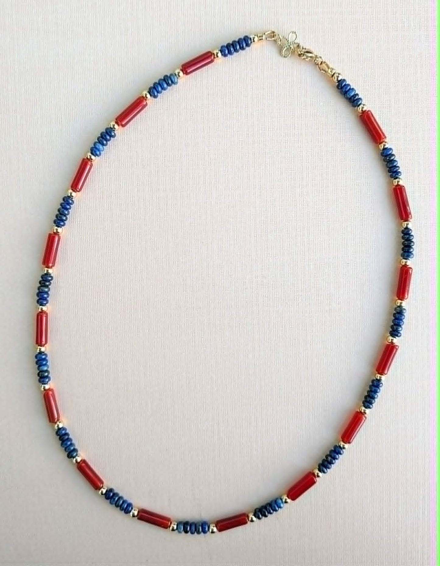 A beaded necklace featuring alternating red cylindrical beads and blue faceted beads separated by small gold spacer rings, displayed on a light beige textured background.
