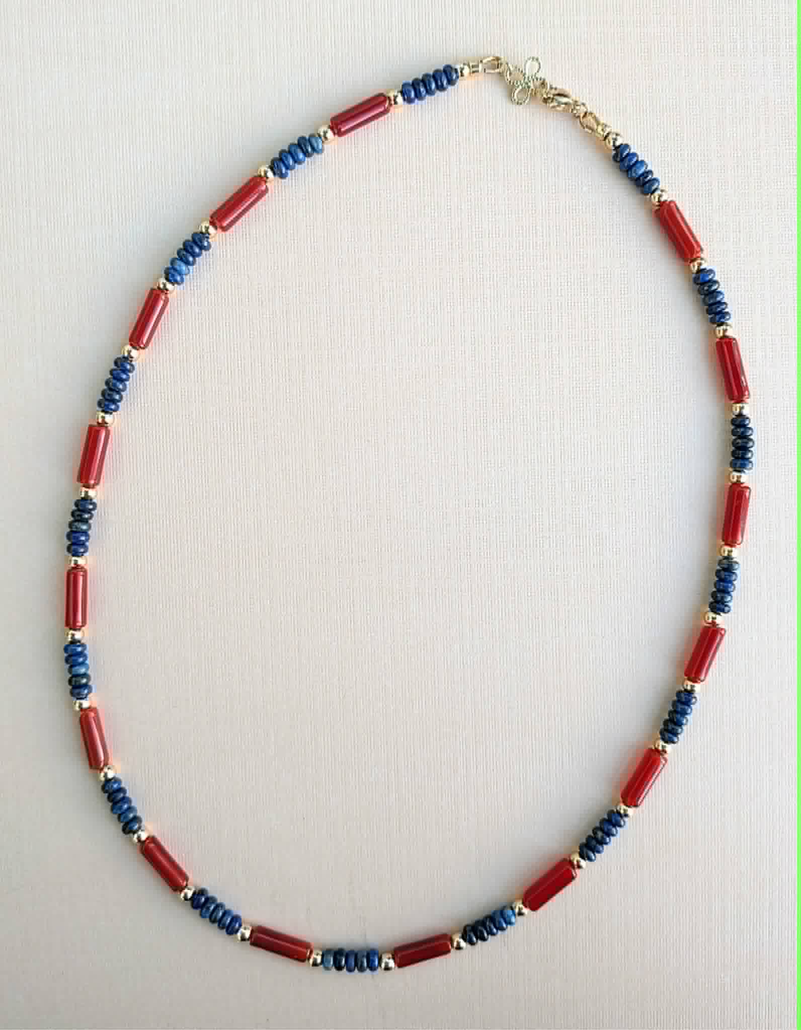 A beaded necklace featuring alternating red cylindrical beads and blue faceted beads separated by small gold spacer rings, displayed on a light beige textured background.