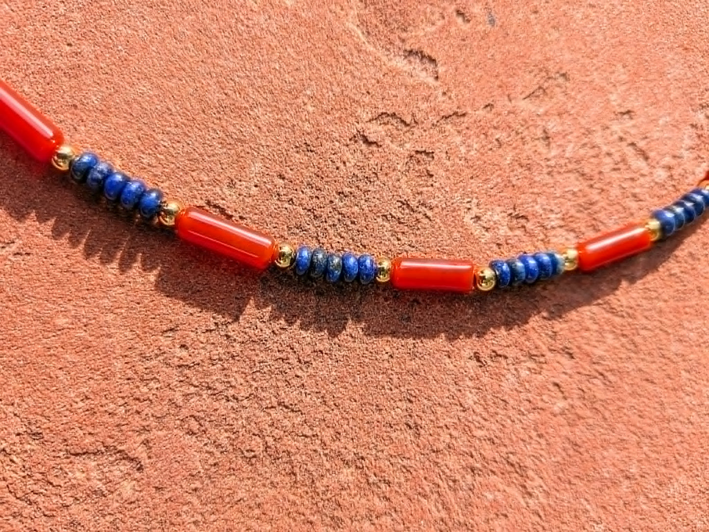 A beaded necklace featuring alternating red carnelian tube beads and blue lapis lazuli beads separated by small gold spacer beads, displayed on a textured terracotta surface.