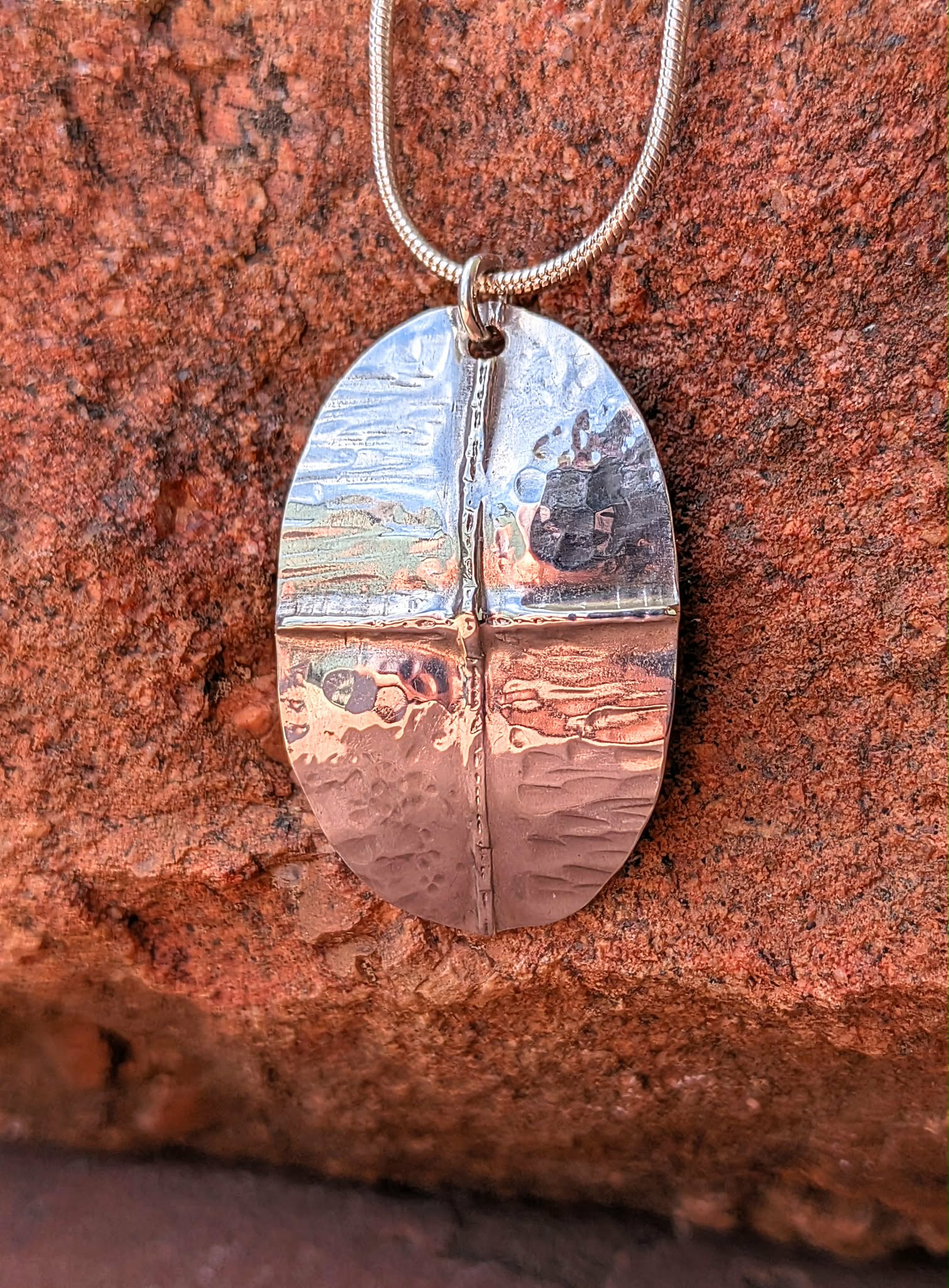 A hammered silver oval pendant necklace resting on a textured red-orange rock surface.