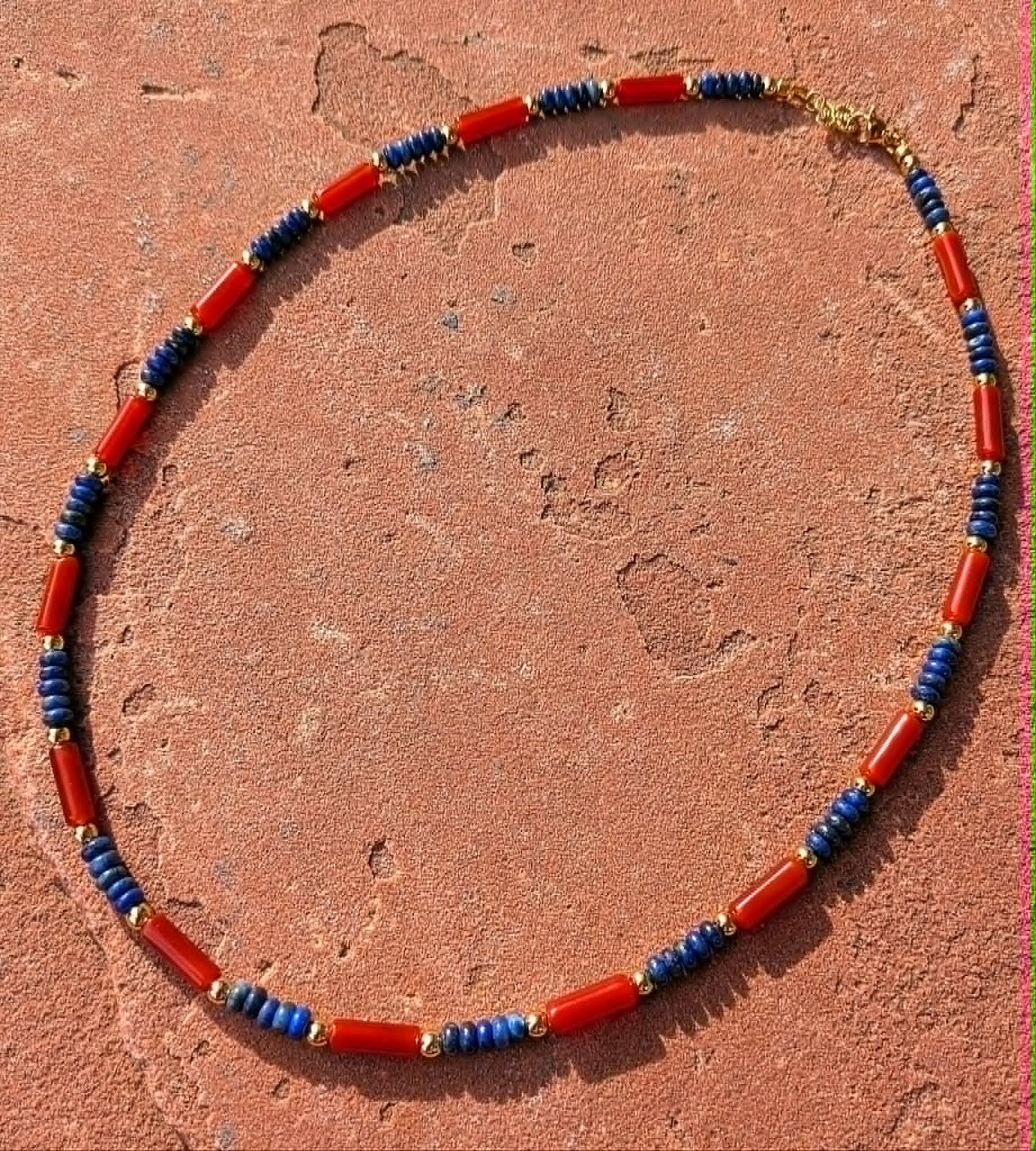 A beaded necklace made of red cylindrical beads separated by blue cylindrical beads and small gold spacer beads, displayed on a textured terracotta surface.