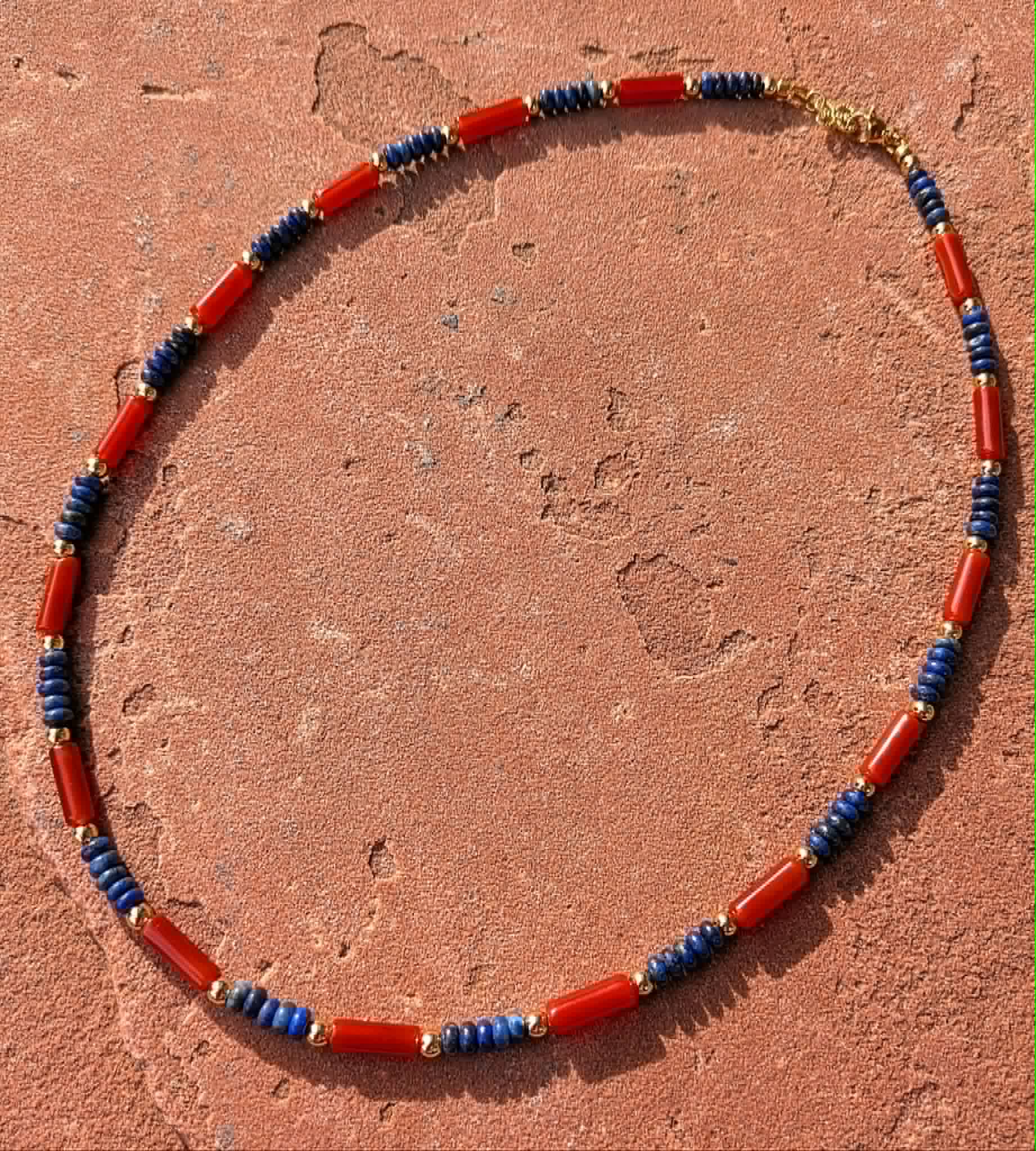 A beaded necklace made of red cylindrical beads separated by blue cylindrical beads and small gold spacer beads, displayed on a textured terracotta surface.
