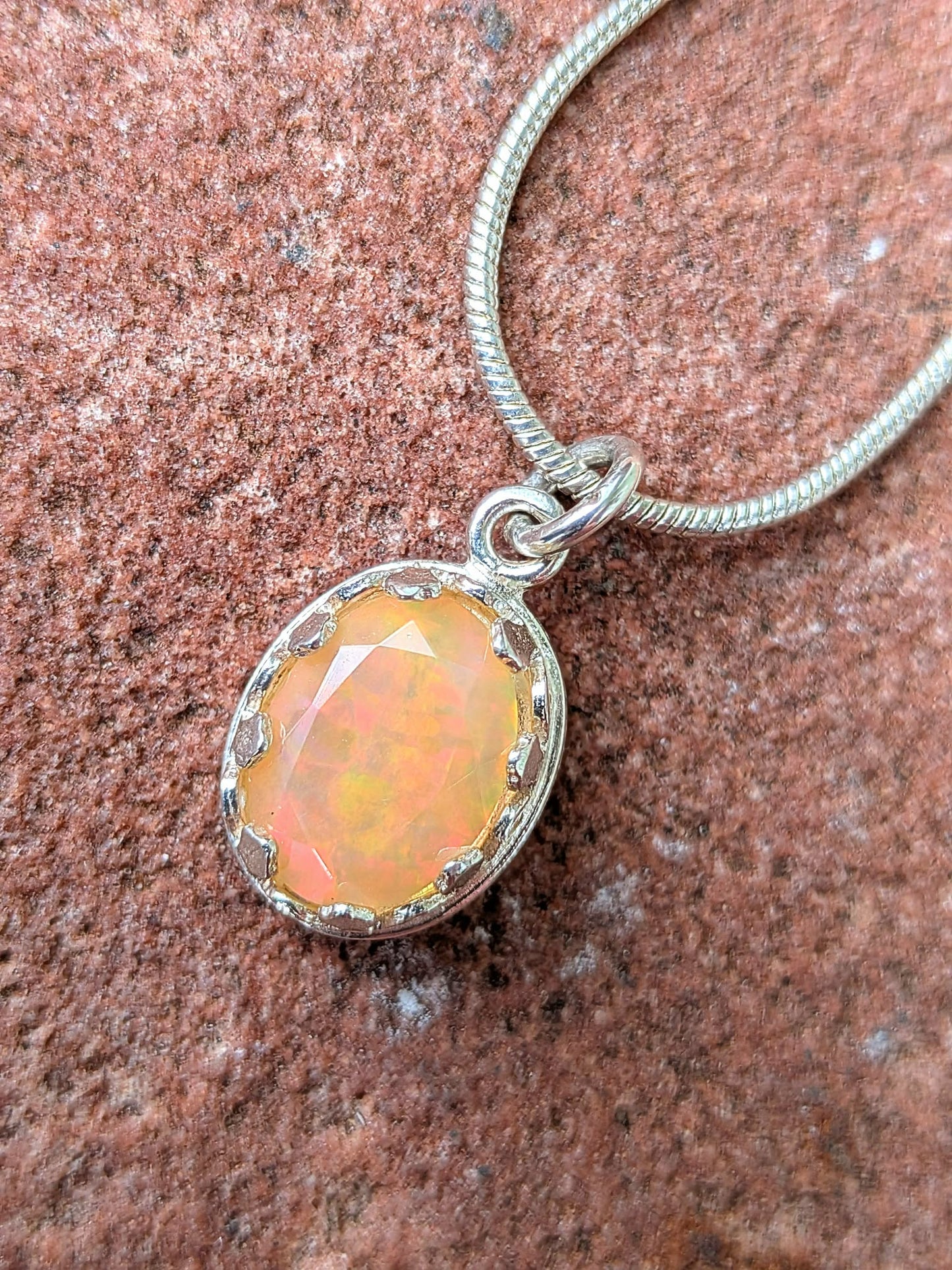An oval pendant necklace featuring a faceted orange opal gemstone in a silver bezel setting, suspended from a silver snake chain and photographed on a textured reddish-brown stone surface.