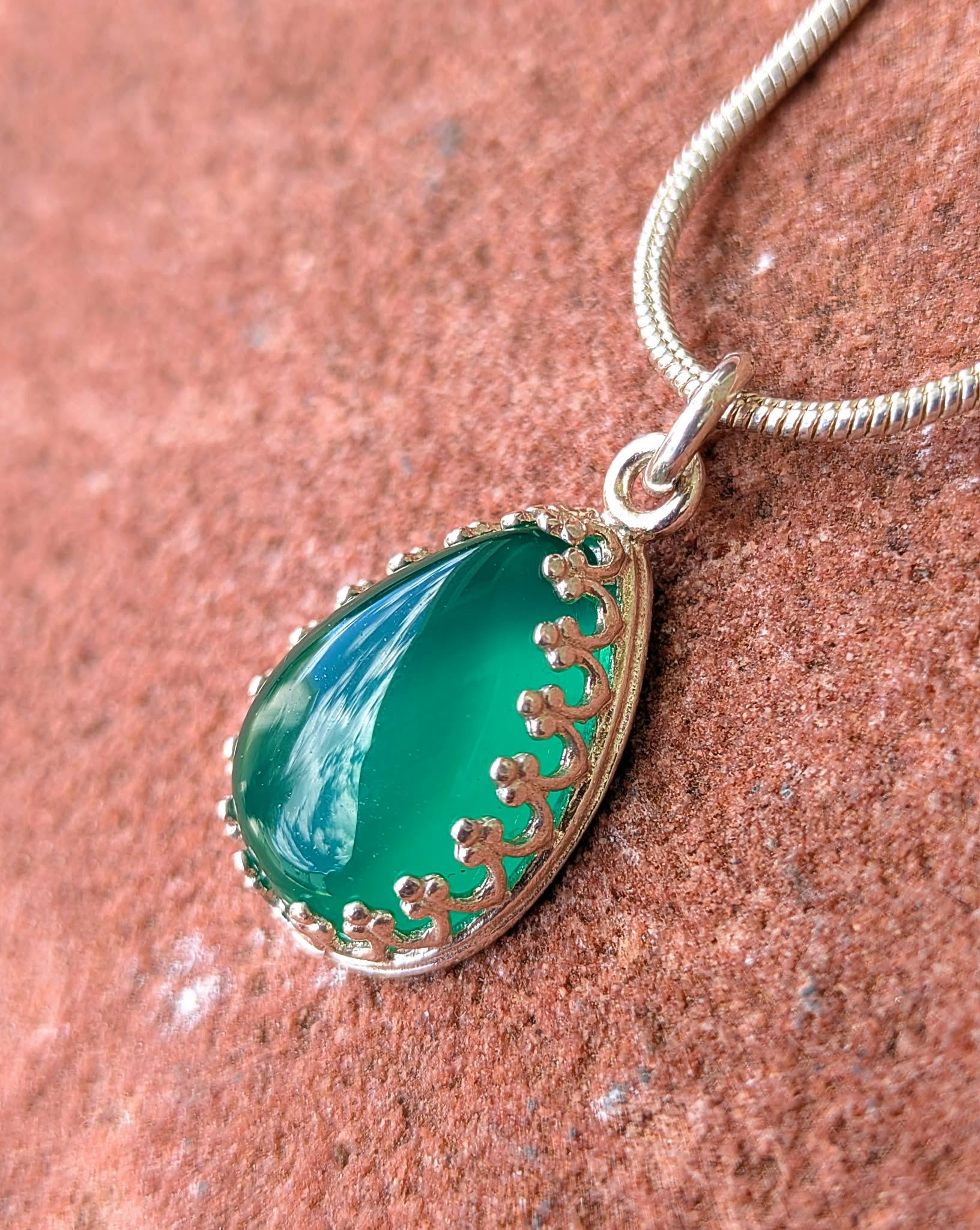 A teardrop-shaped green stone pendant with a silver filigree border and a silver snake chain, resting on a textured reddish-brown stone surface.