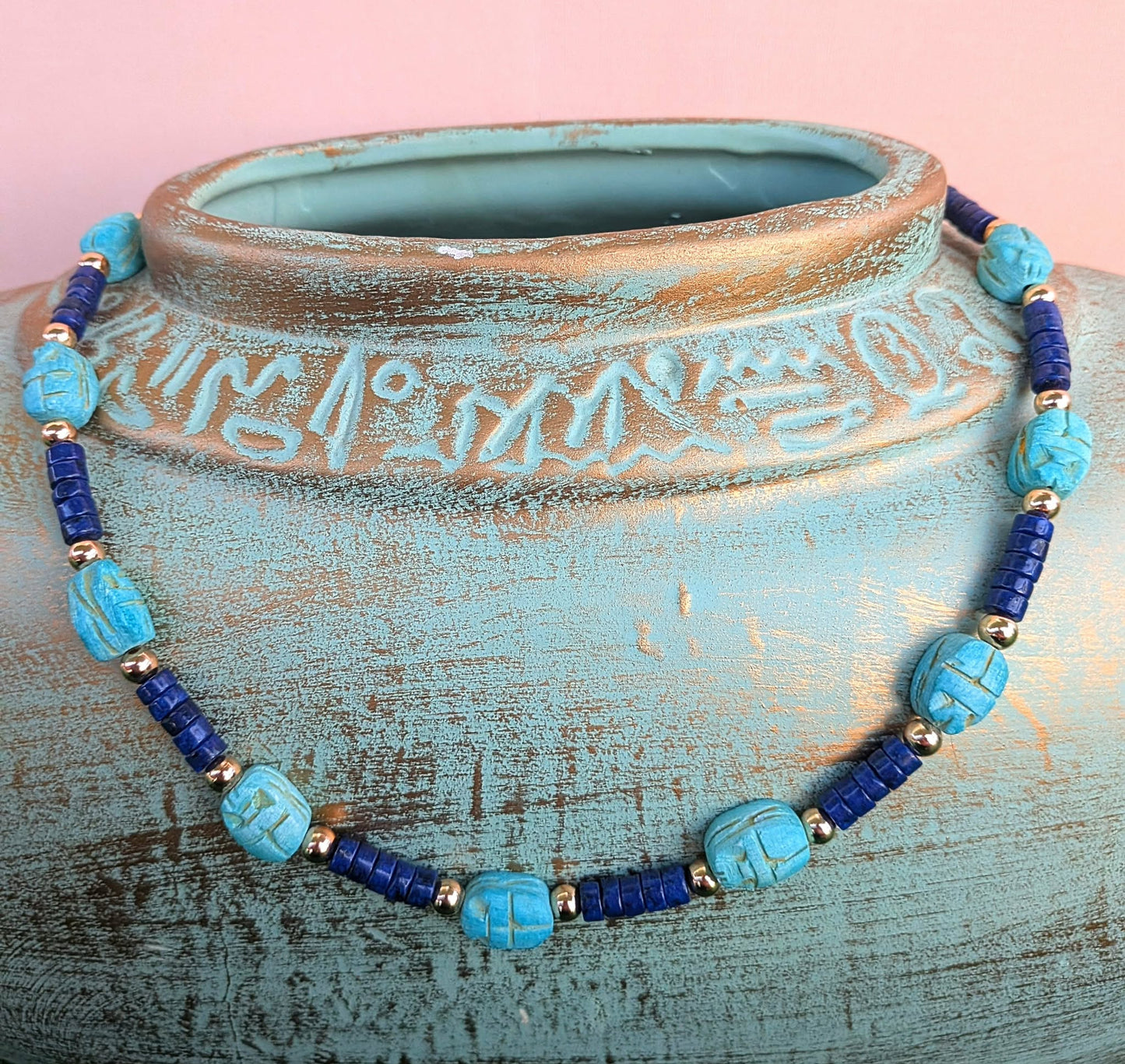 A beaded necklace with turquoise and dark blue beads, accented by small gold spacer beads, displayed on a distressed turquoise ceramic vase against a pink background.
