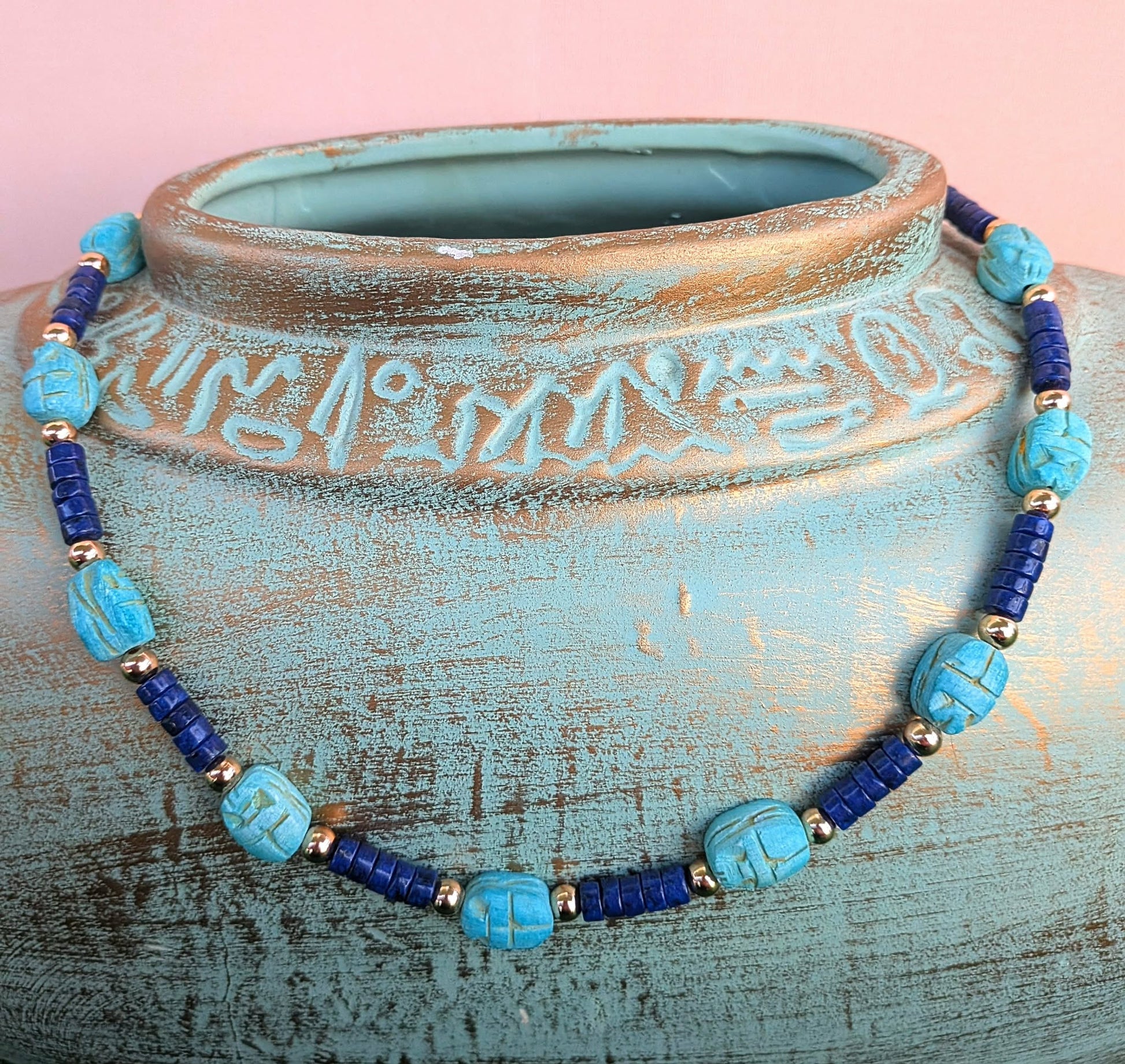 A beaded necklace with turquoise and dark blue beads, accented by small gold spacer beads, displayed on a distressed turquoise ceramic vase against a pink background.
