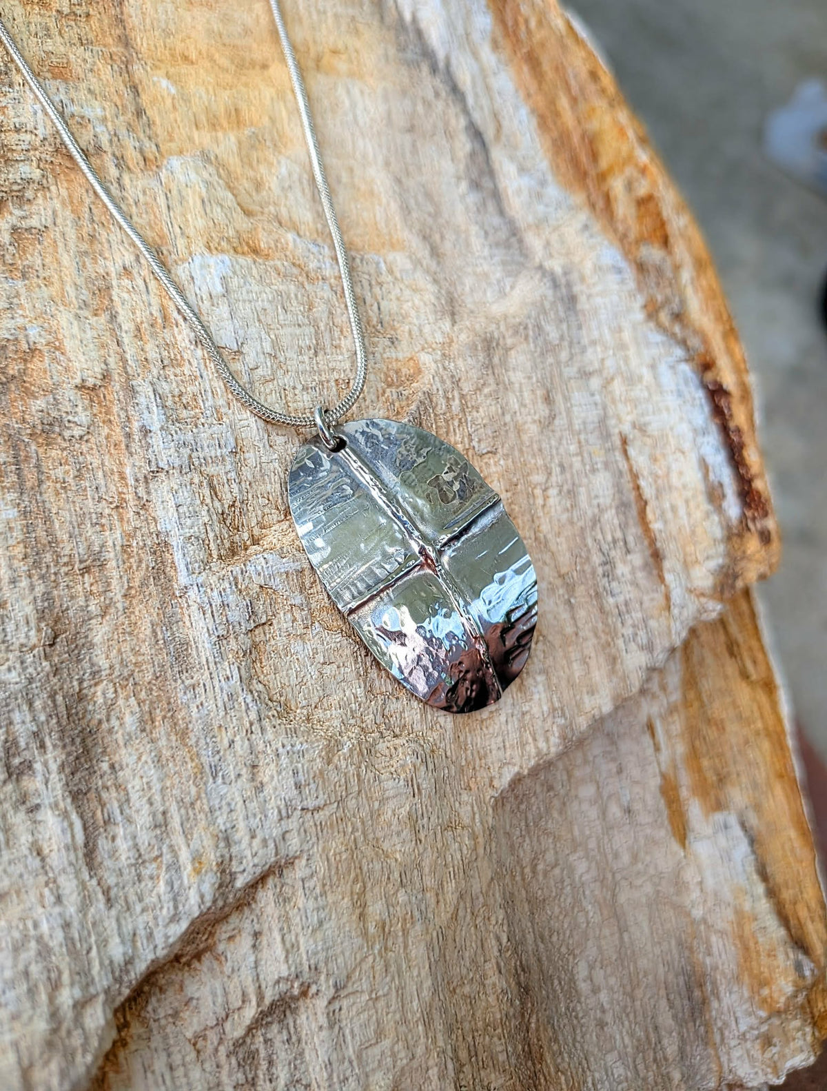A silver metal pendant necklace with a reflective, shield-like polished surface, resting on a textured piece of wood.