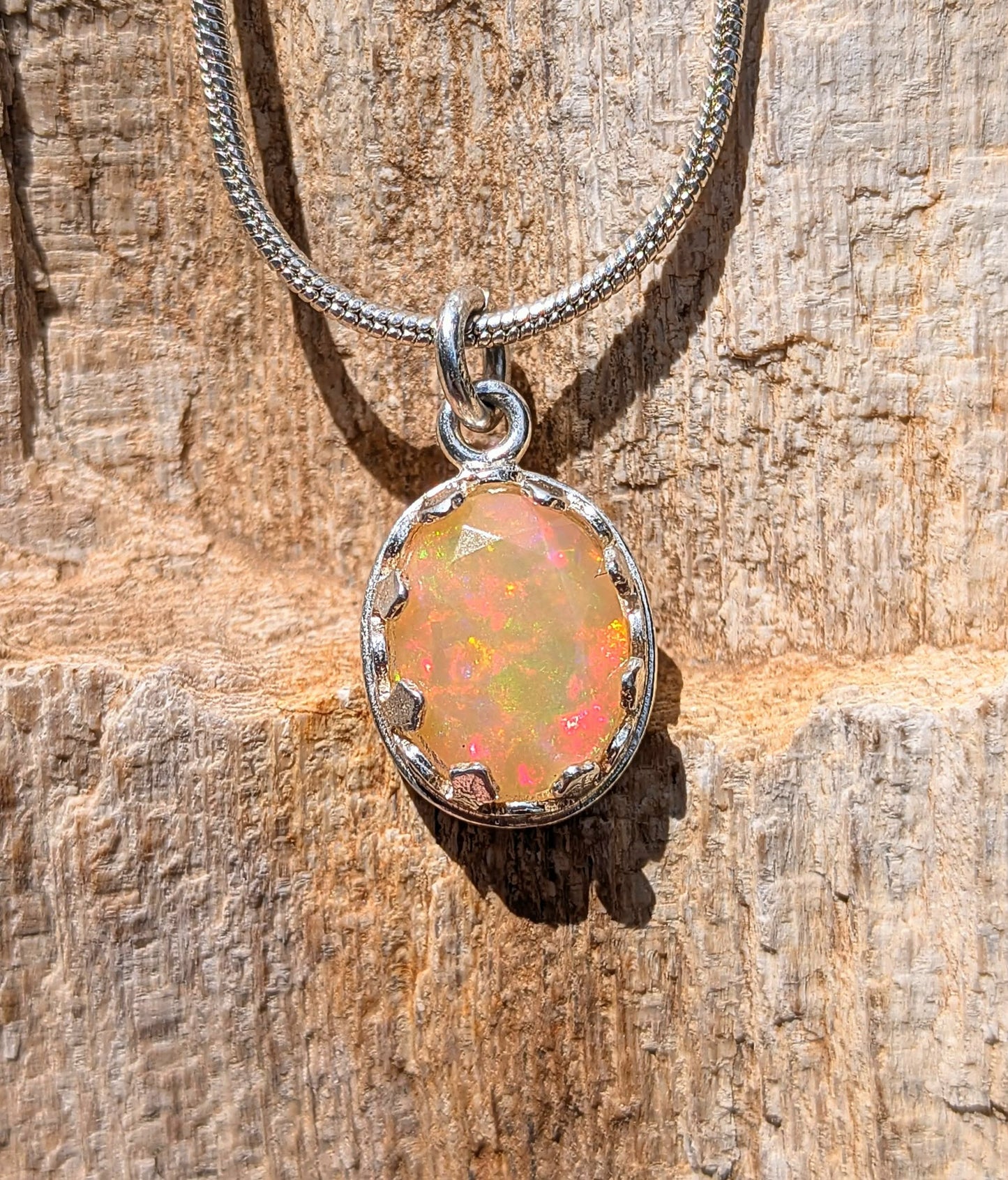 A silver chain necklace with an oval pendant featuring a colorful opal gemstone, displayed on a textured wooden surface.