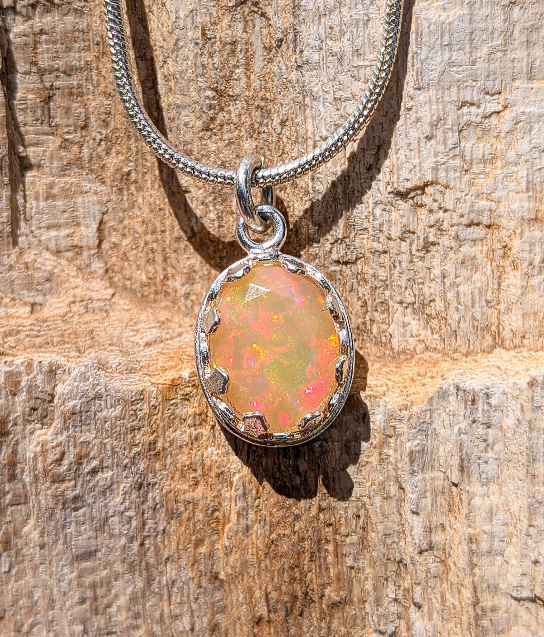 A silver chain necklace with an oval pendant featuring a colorful opal gemstone, displayed on a textured wooden surface.