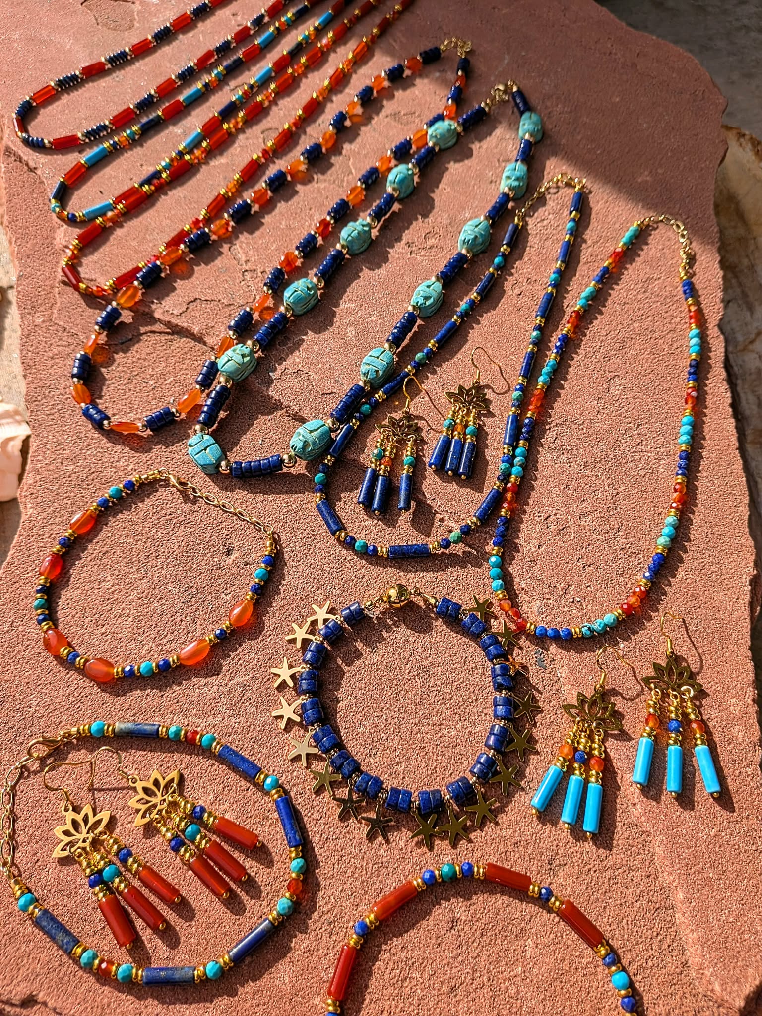 A collection of handmade beaded jewelry, including necklaces, bracelets, and earrings, displayed on a textured terracotta surface. The jewelry features vibrant blue, orange, and turquoise beads with gold-toned metal charms and tassels.