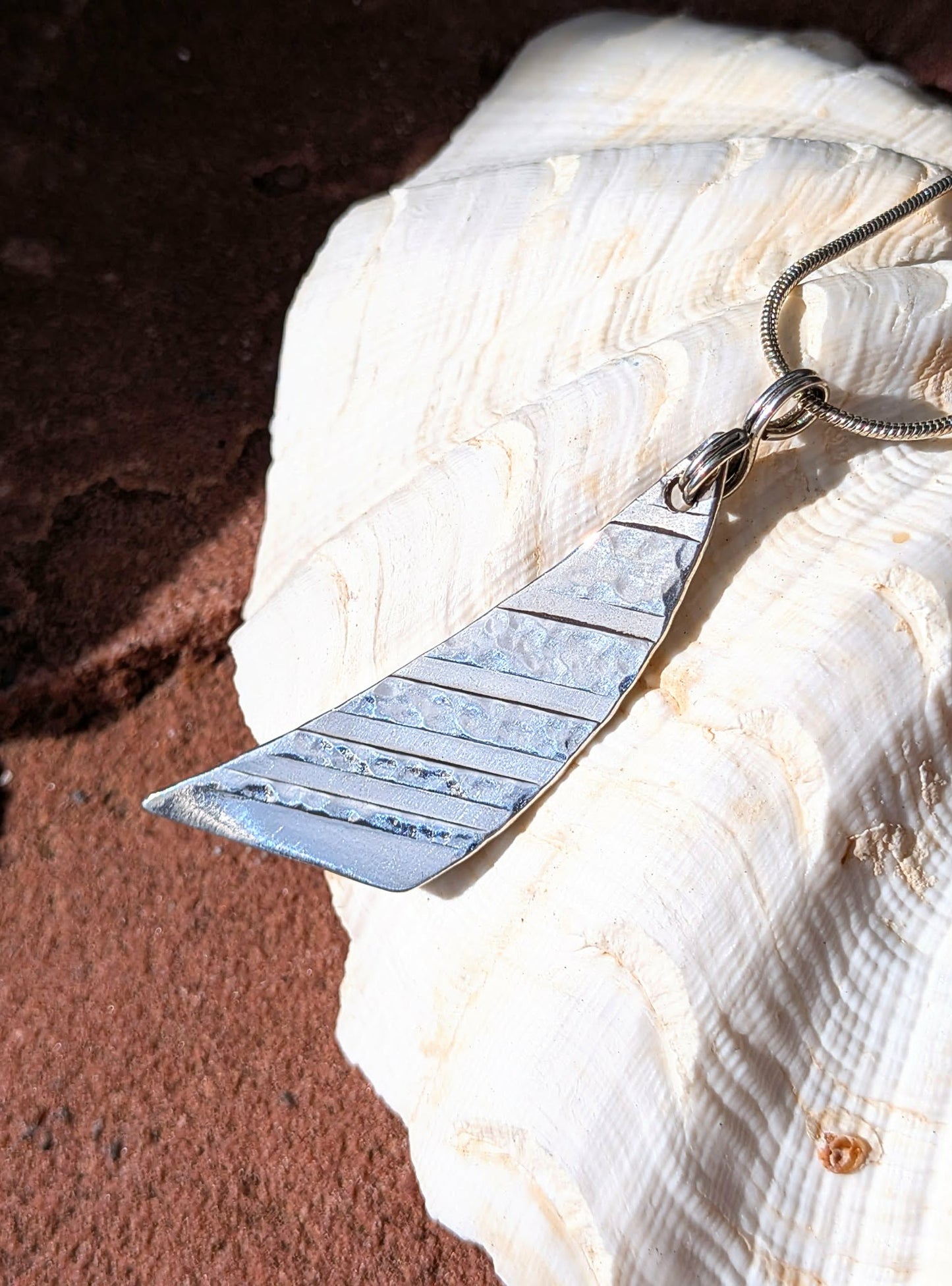 A hammered silver pendant necklace with a geometric, sail-like triangular shape, resting on a large white seashell against a dark brown textured background.