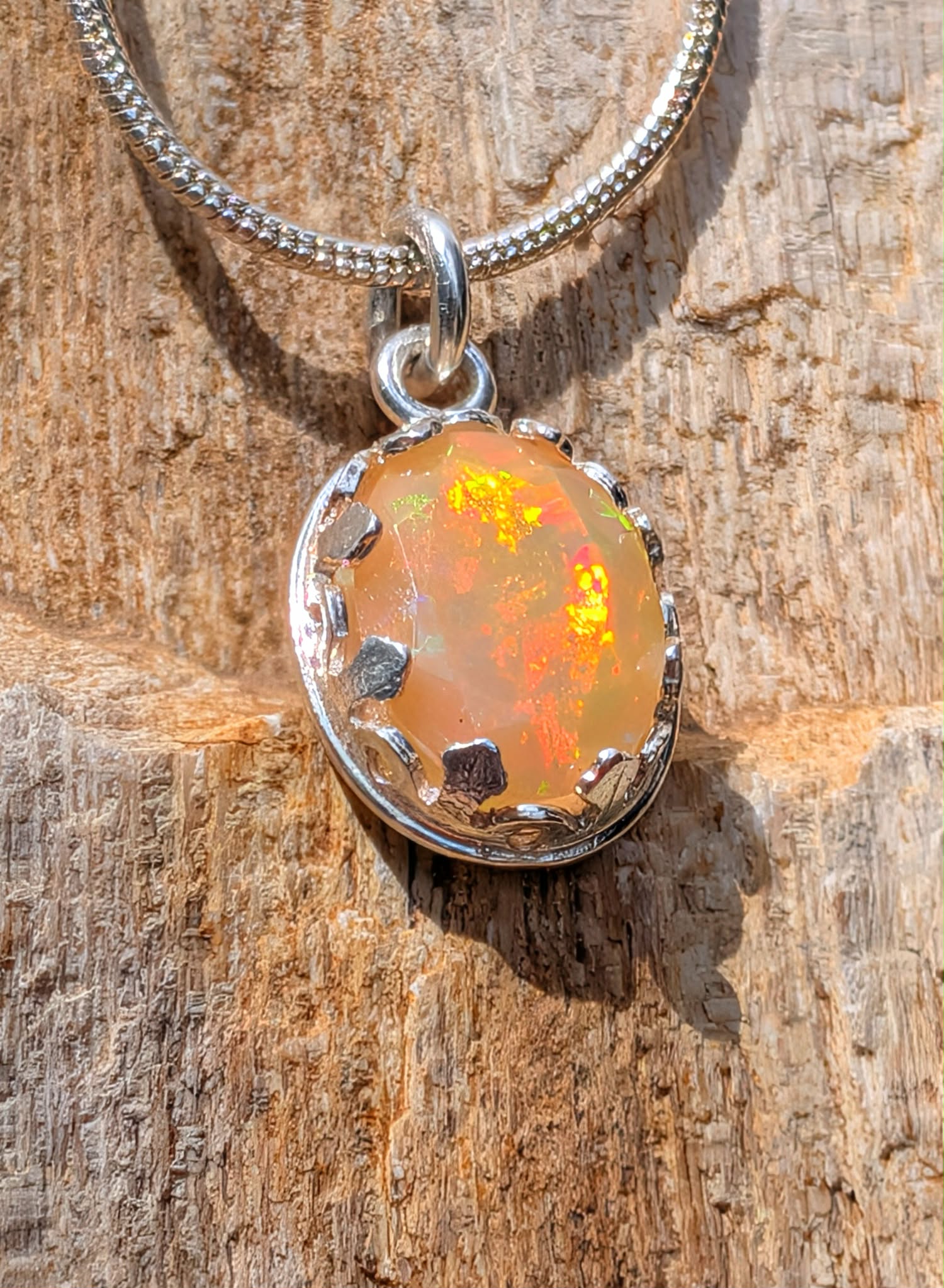 A silver chain necklace with an oval pendant featuring a warm orange and yellow opal-like gemstone in a textured silver bezel, displayed on a rustic wood surface.