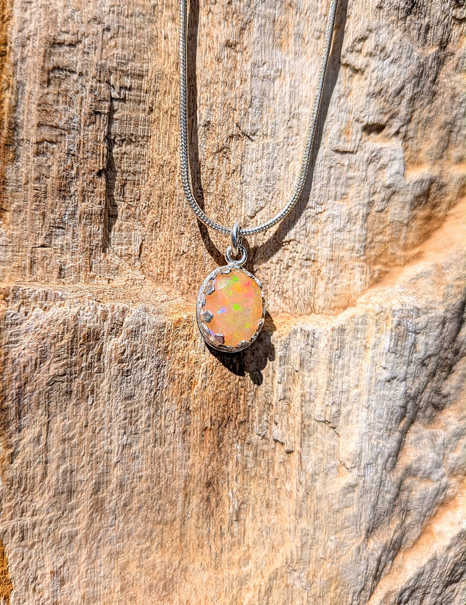 A silver snake chain necklace with an oval opal pendant, displayed against a textured natural rock background.