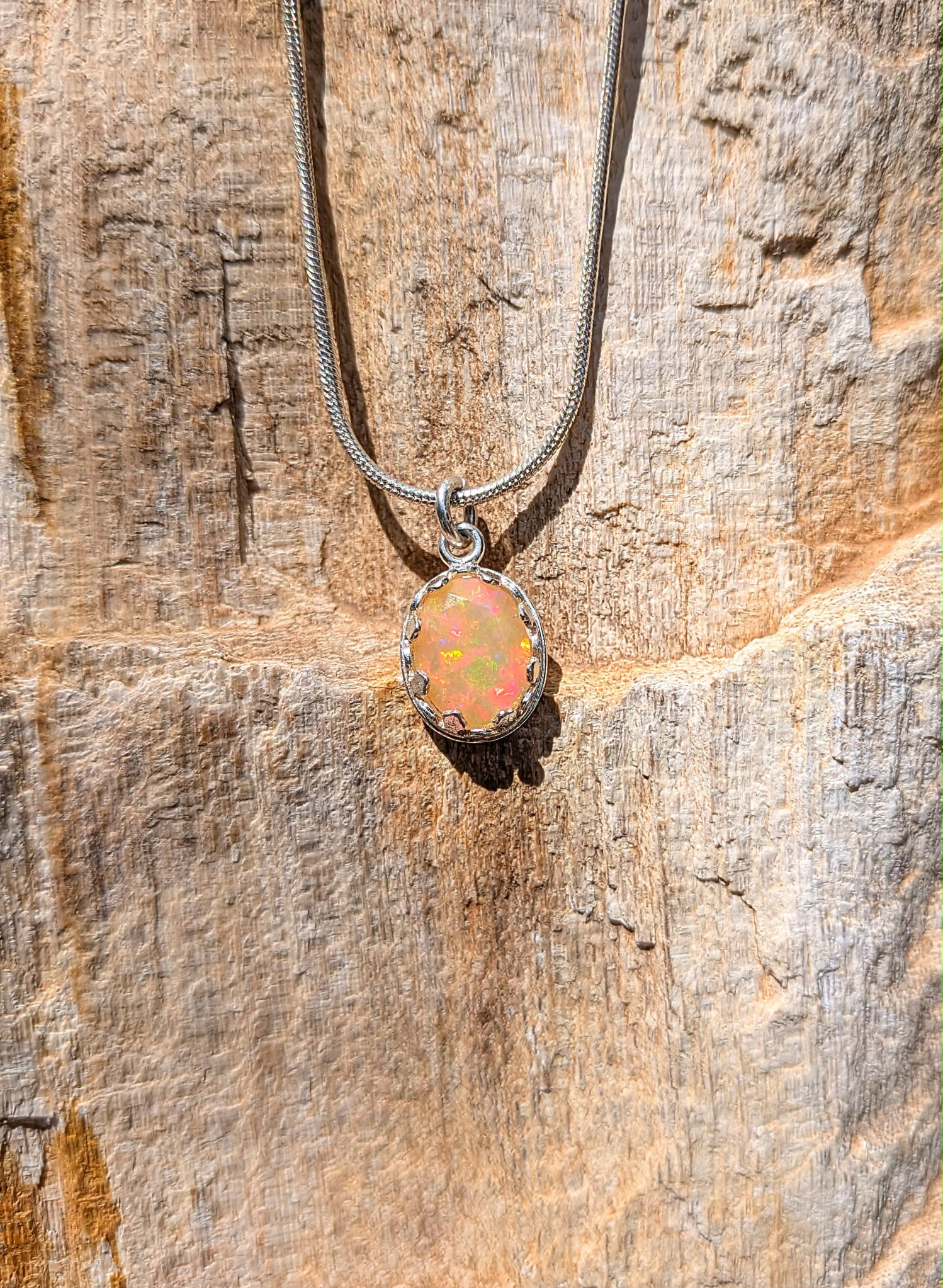 A silver snake chain necklace with an oval opal pendant, displayed against a textured natural wood background.