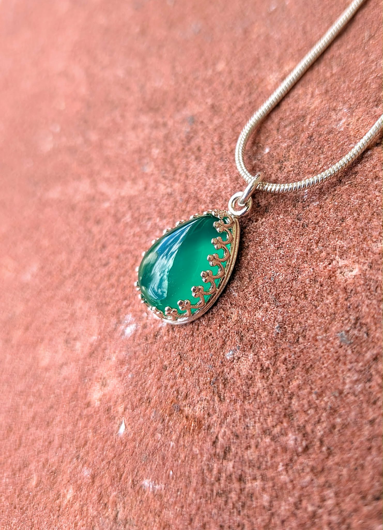A teardrop-shaped green stone pendant with a gold-toned floral border, hanging from a silver snake chain against a textured reddish-brown stone surface.