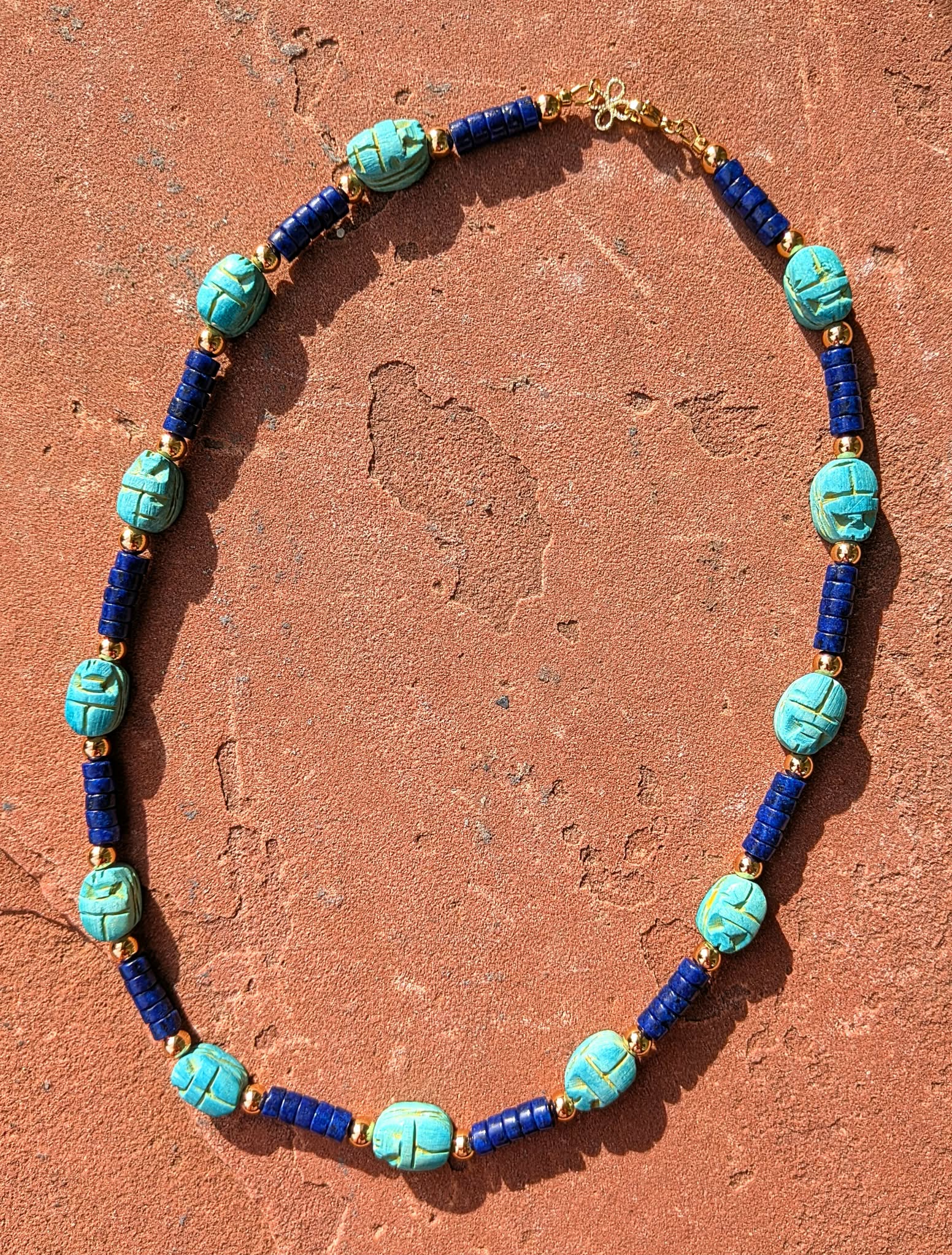A beaded necklace with turquoise scarab beetle beads, dark blue cylindrical beads, and small gold spacer beads, arranged in an oval on a textured terracotta surface.