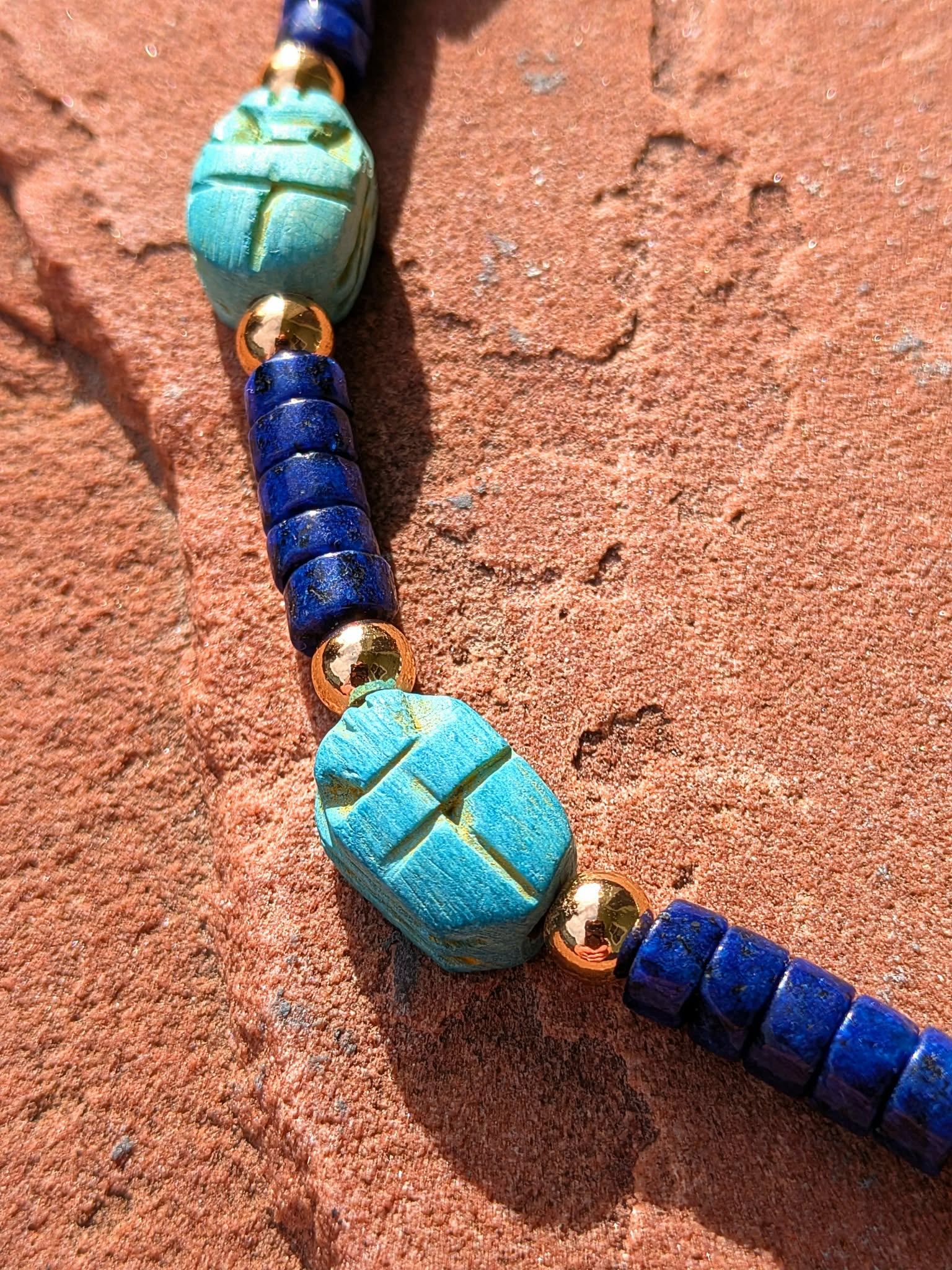 A close-up of a beaded necklace featuring turquoise scarab beetle pendants, deep blue cylindrical beads, and small gold spacer beads, laid on a textured reddish-brown stone surface.