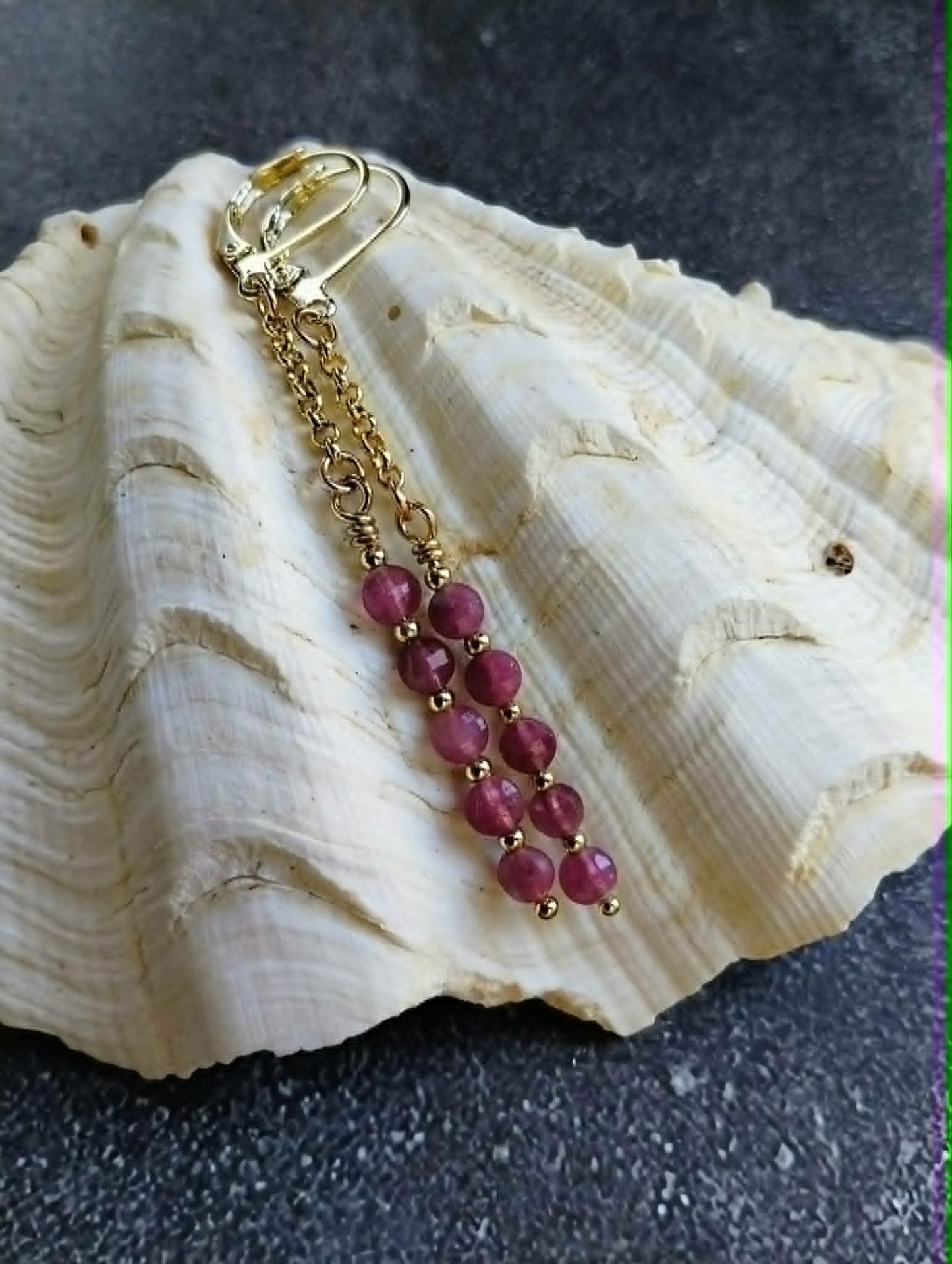 A gold-toned dangle earring with a lobster clasp and a series of round faceted pinkish-purple beads, displayed on a large cream-colored seashell against a dark textured background.