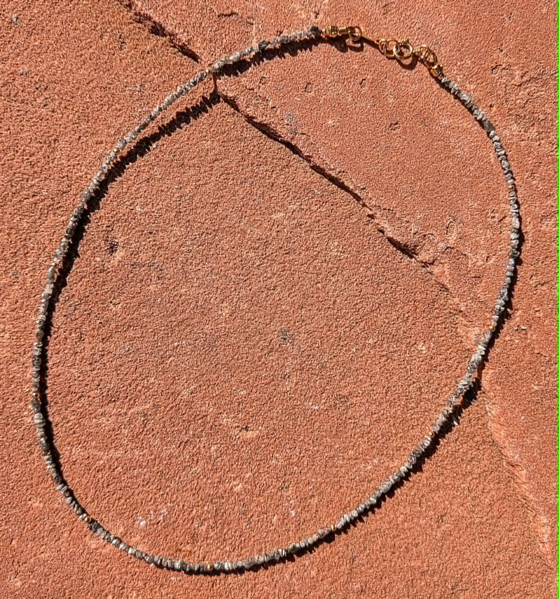 A beaded necklace made of small gray and white diamond uncut nugget beads with a gold clasp, laid flat on a textured terracotta surface.