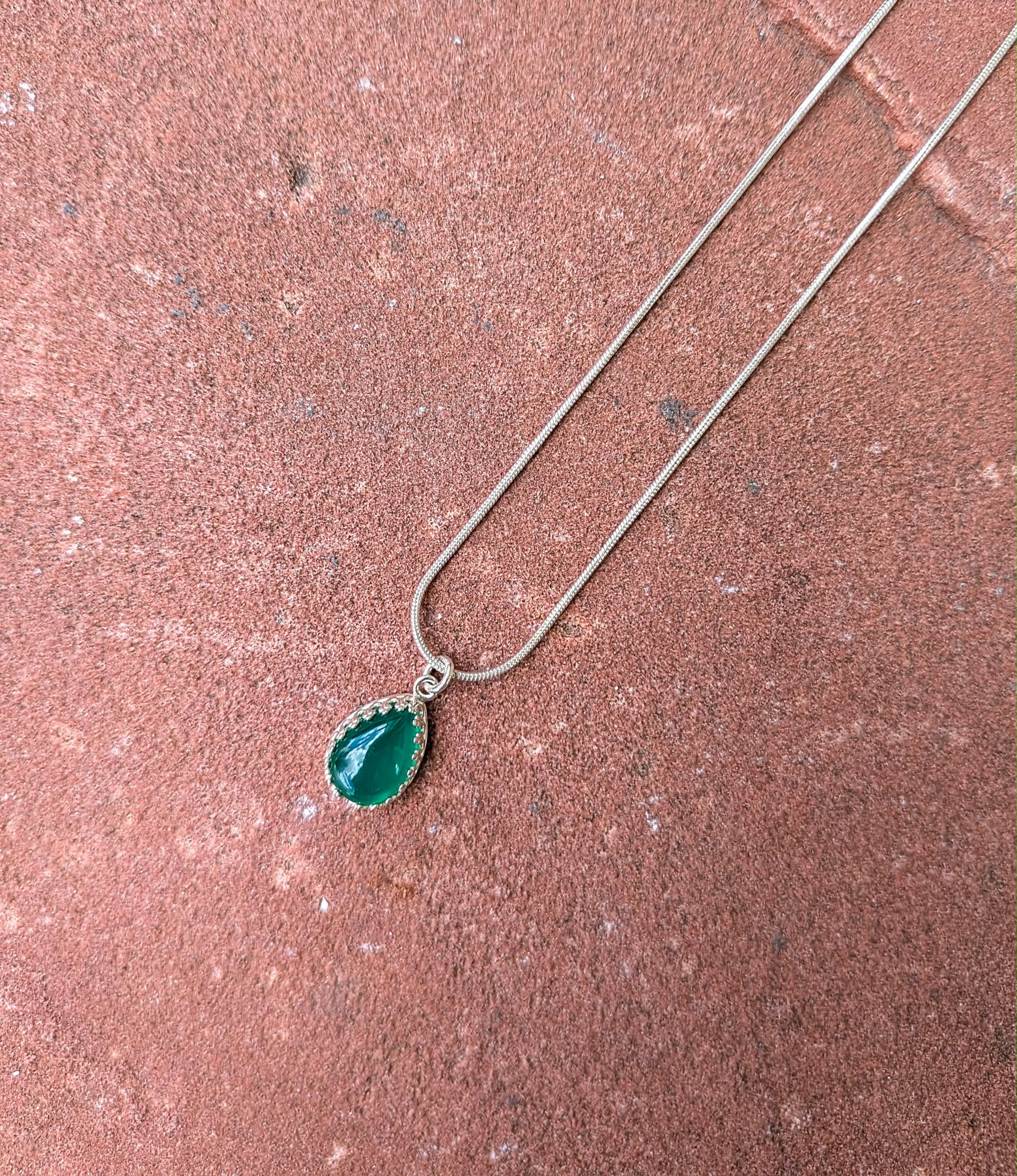 A silver-tone chain necklace with a teardrop-shaped green gemstone pendant, laid diagonally on a textured reddish-brown stone surface.
