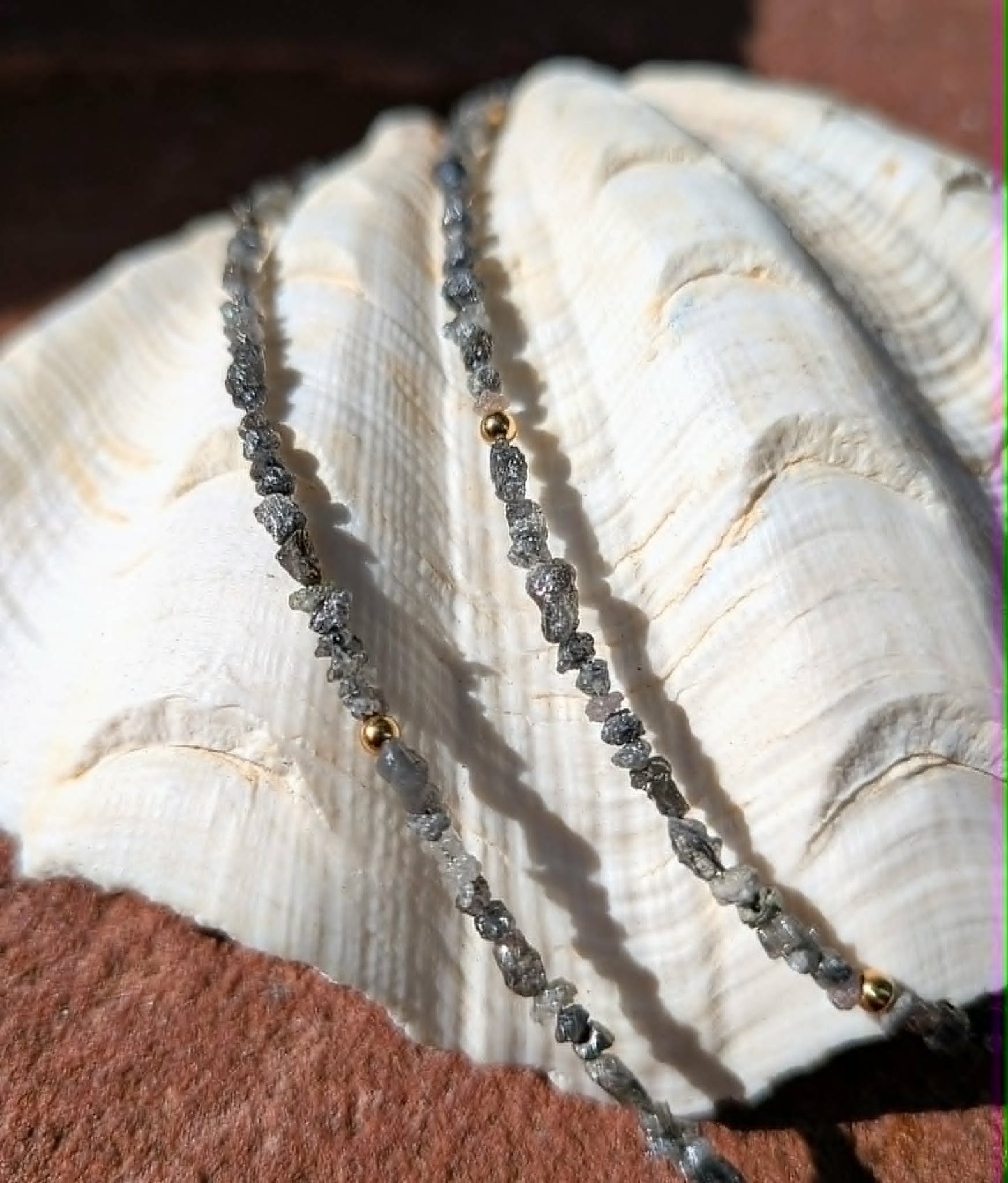 A close-up of a beaded strand jewelry piece draped over a large white seashell, resting on a reddish-brown textured surface. The beads are primarily dark gray rough uncut diamond nugget beads with small gold spacer beads.