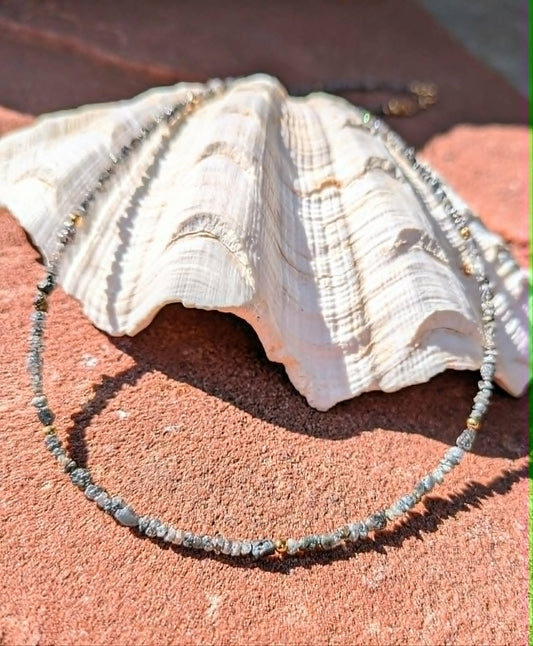 A beaded necklace made of small gray and white uncut rough nugget diamond beads, with tiny gold spacer beads, draped around a large white seashell on a textured reddish-brown surface.