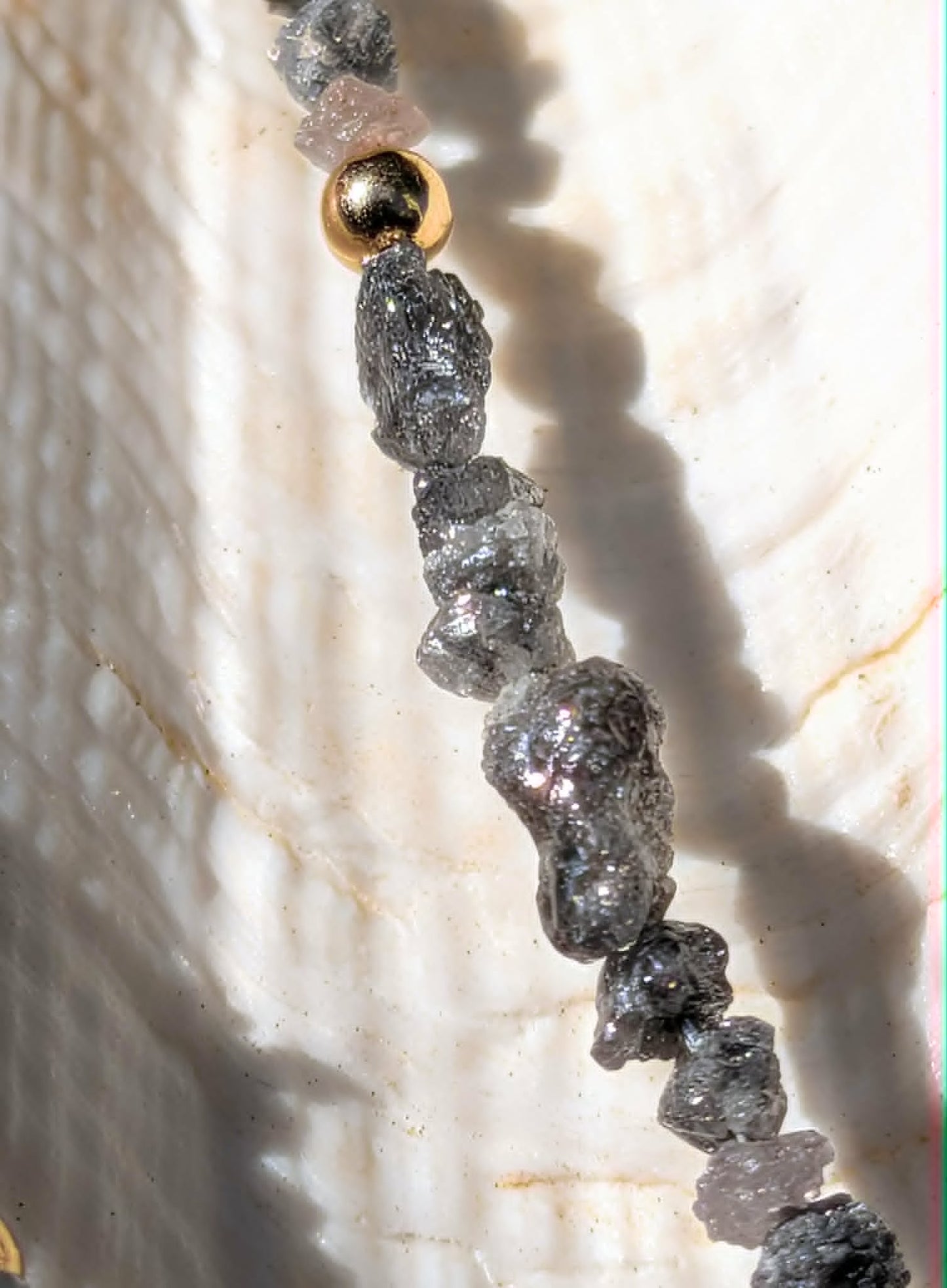 A close-up of a beaded strand featuring rough, dark gray to black rough uncut diamond nugget beads and a single metallic gold bead, resting on a textured white clam shell.