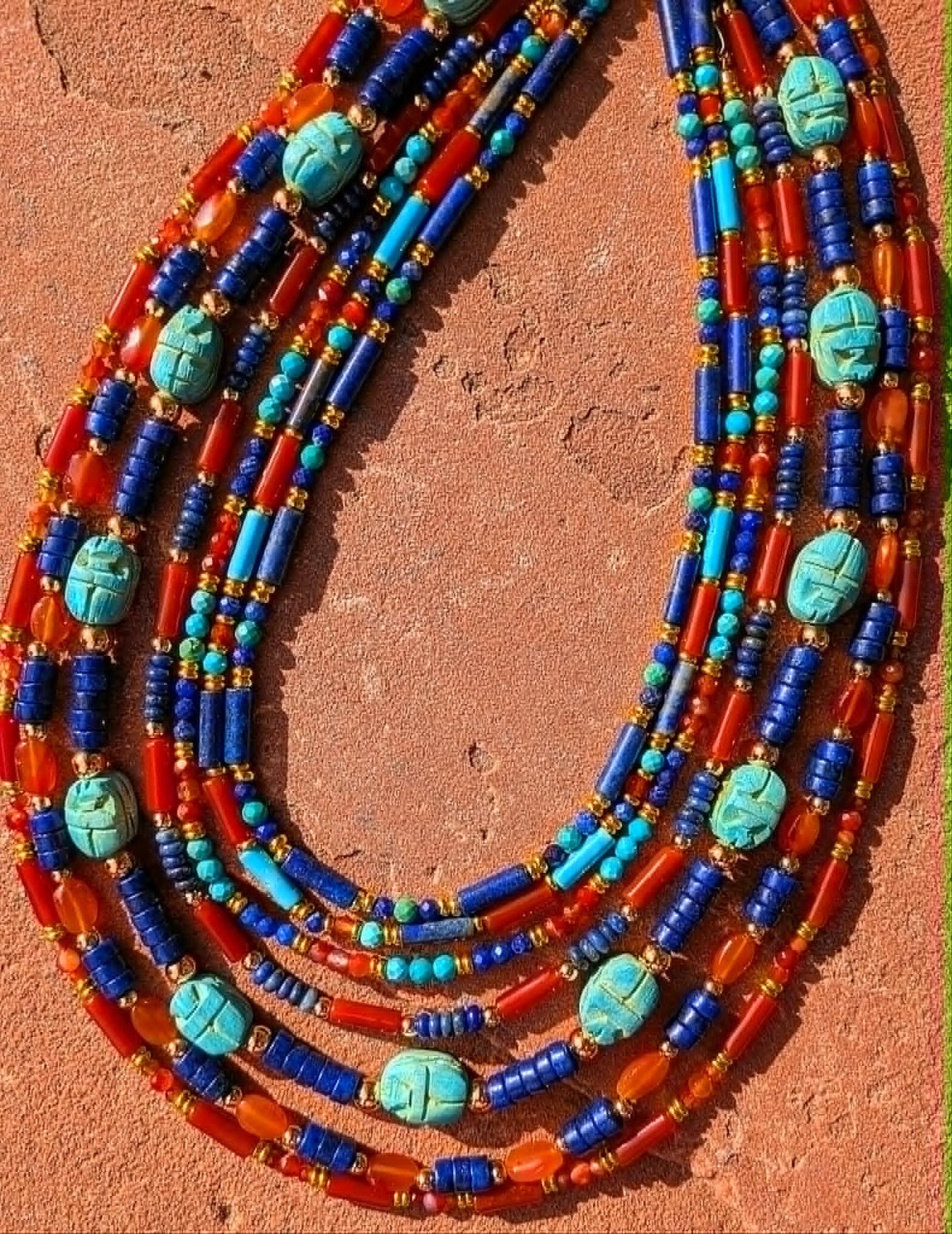 A multi-strand beaded necklace laid flat on a textured terracotta surface. The necklace features alternating strands of bright blue beads, orange-red beads, turquoise blue beads, and carved turquoise oval pendants with a central black line detail.