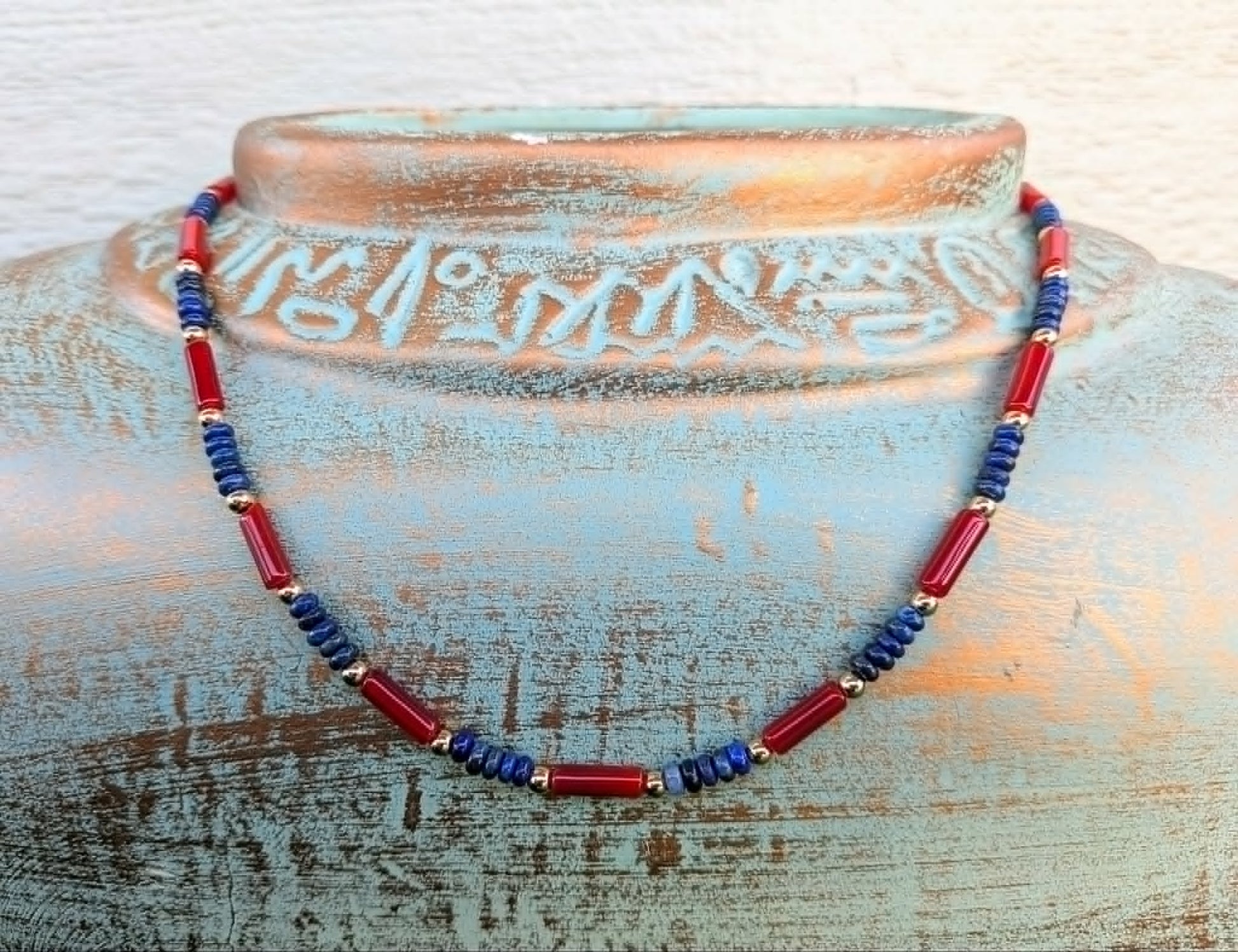 A beaded necklace with alternating red and blue cylindrical beads separated by small gold spacer rings, displayed draped around a rustic distressed blue ceramic vase.