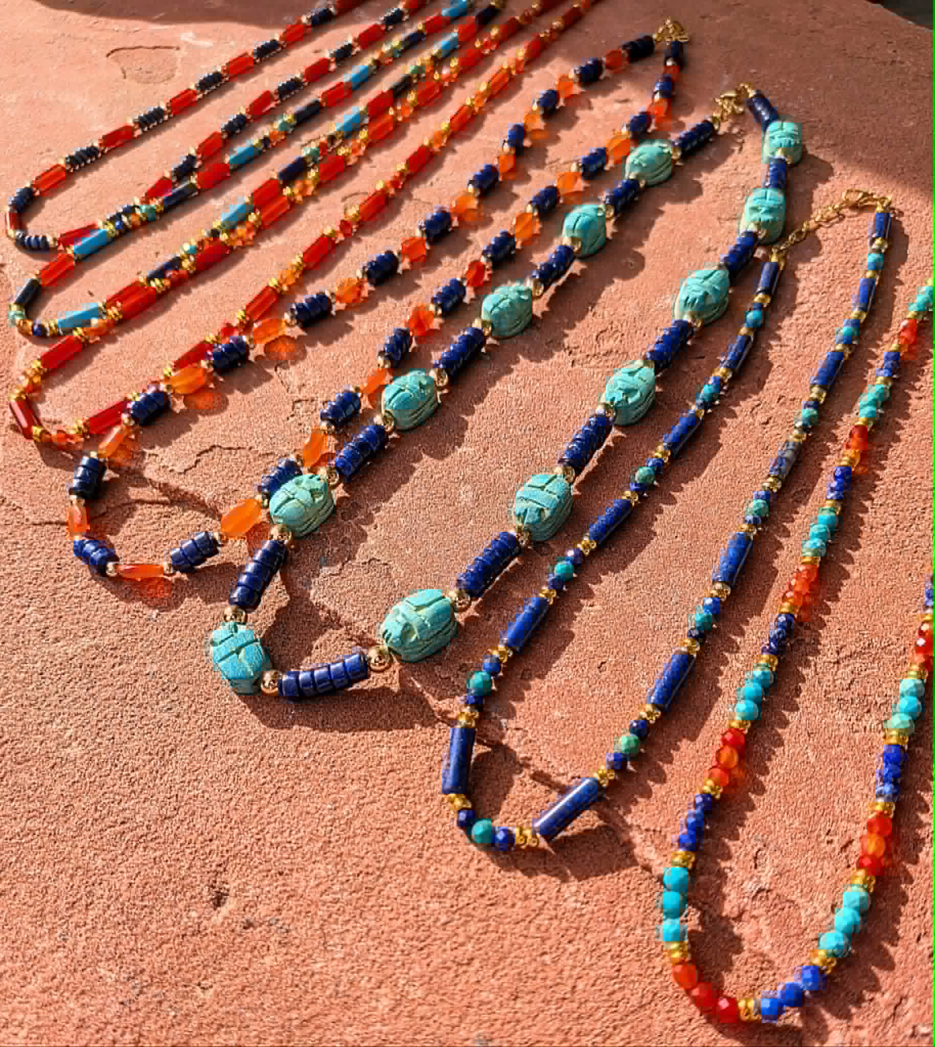 Several colorful beaded necklaces are arranged diagonally on a textured terracotta surface. The necklaces feature turquoise, royal blue, orange, and red beads with small gold-toned spacer details.