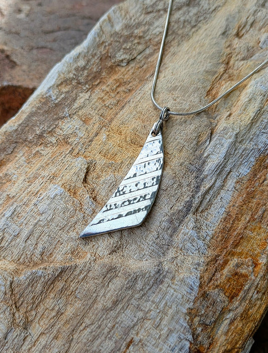 A silver geometric pendant necklace with a textured, hammered triangular design, resting on a rustic, textured stone surface.