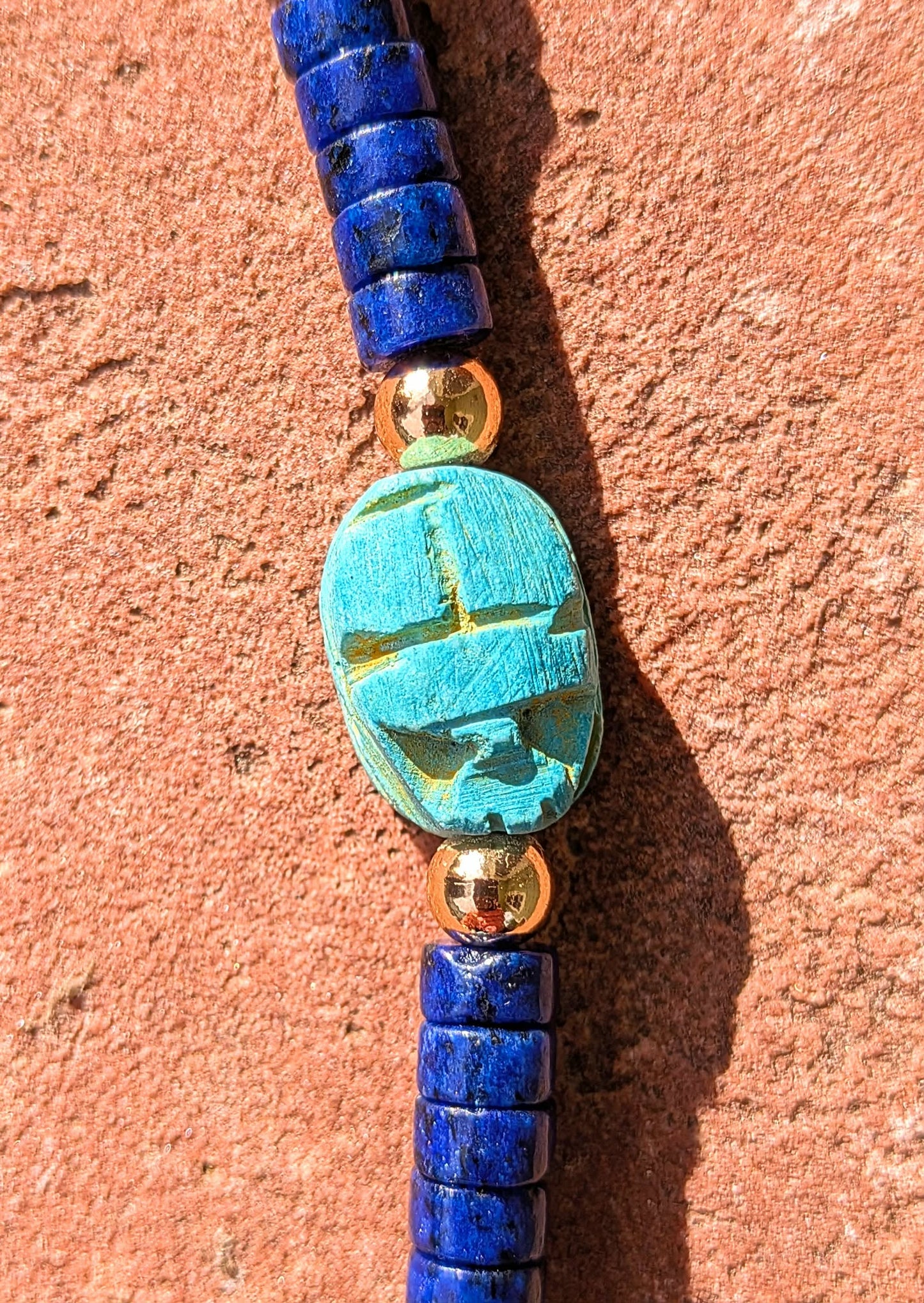 A beaded necklace featuring deep blue cylindrical beads, a central turquoise cabochon with a cross motif, and small gold spacer beads, displayed against a textured terracotta background.