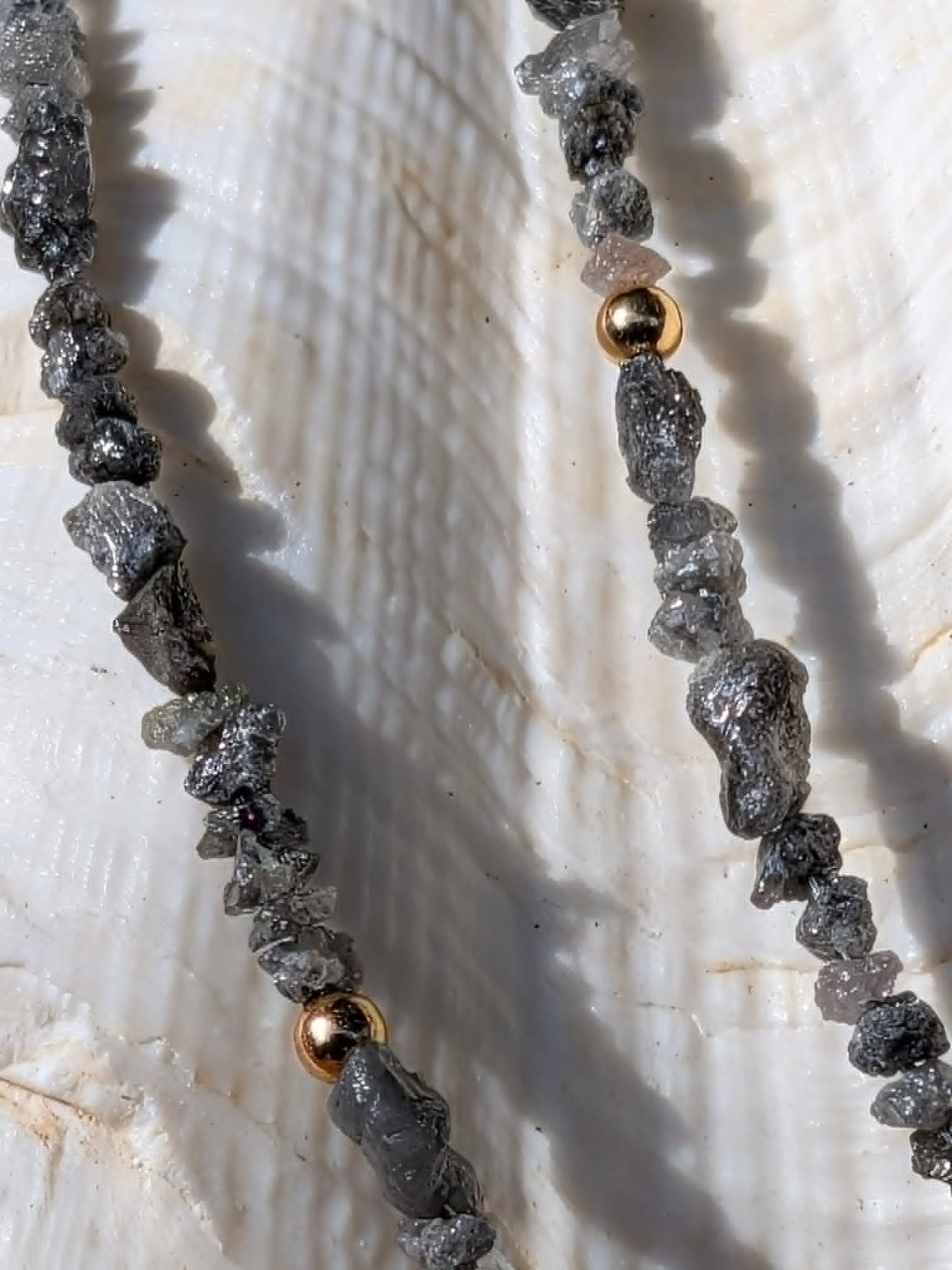 A close-up of a beaded necklace made of rough dark gray uncut rough diamond nugget beads with small gold spacer beads, laid on a white seashell.