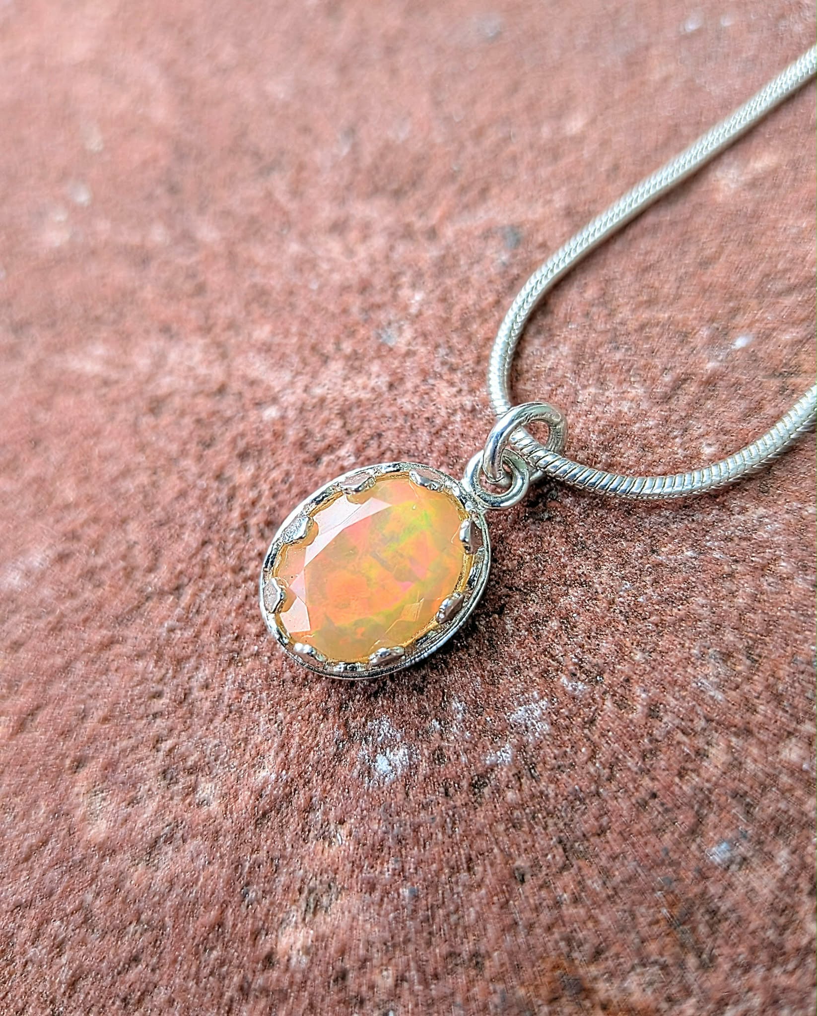 A silver snake chain necklace with an oval bezel-set opal pendant, displayed on a textured reddish-brown stone surface.