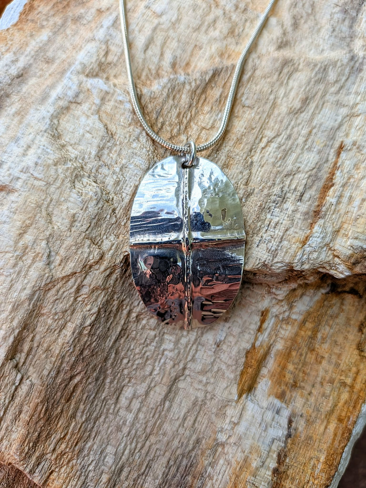 A silver oval pendant necklace with a textured, hammered finish and a central vertical seam, resting on a textured natural wood background.